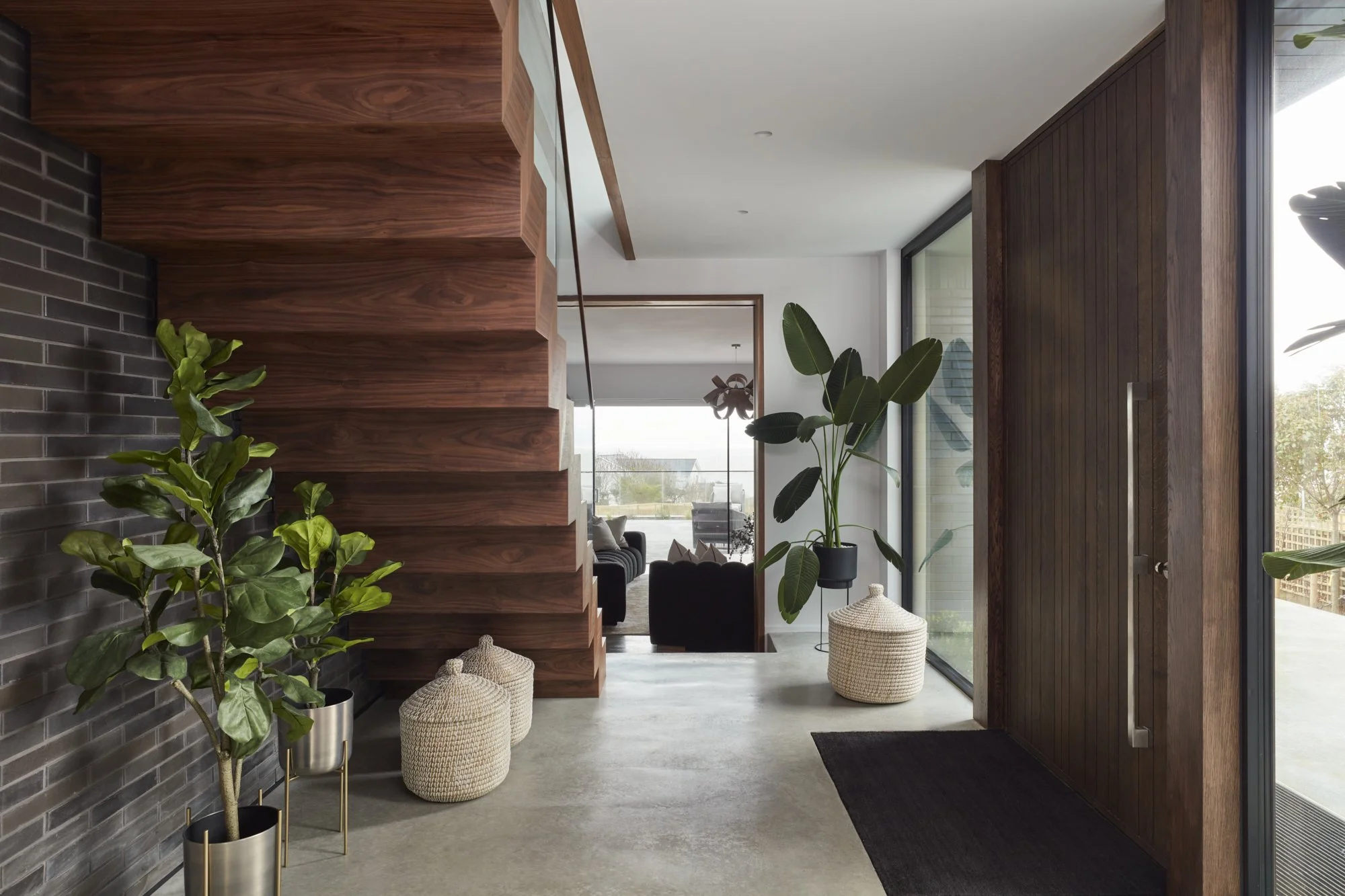Modern interior with dark brick wall, wooden staircase, green plants in metallic pots, wicker baskets, large windows, and a view of outdoor landscape.