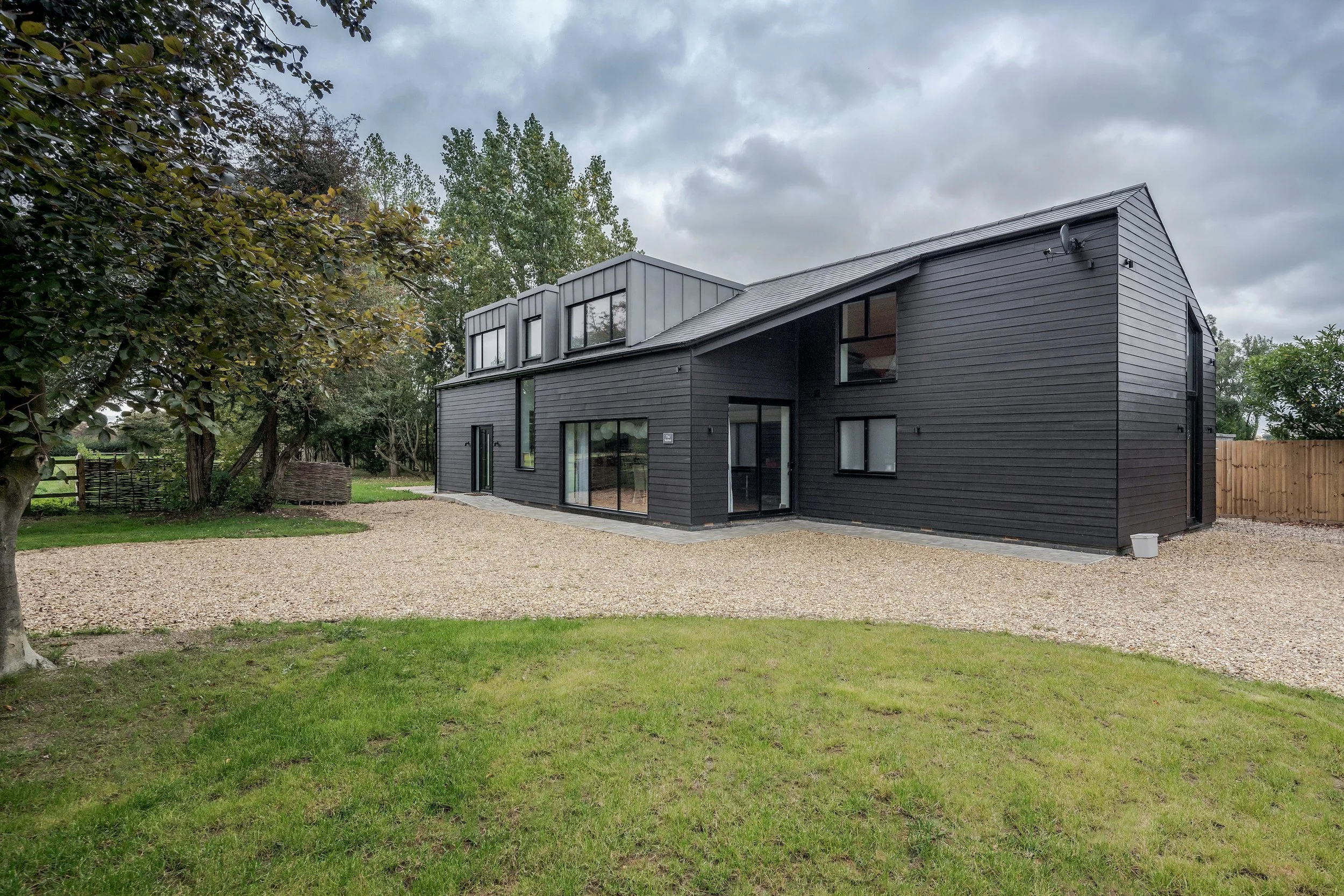 Modern black house with large windows, surrounded by trees and a gravel yard, under a cloudy sky.
