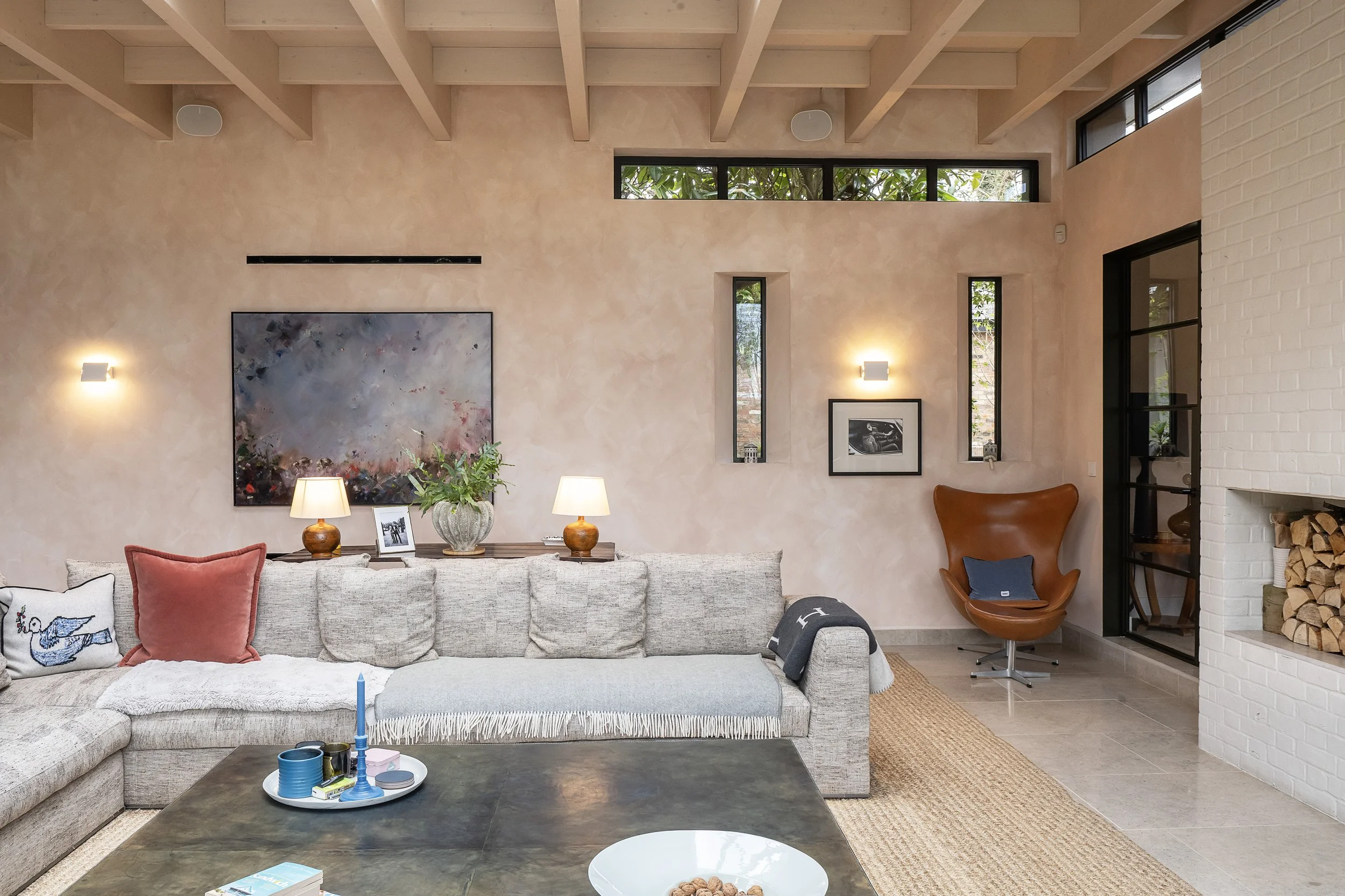 Living room with beige sofa, decorative pillows, a black coffee table with items, a side table with lamps, wall art, a leather armchair, and a white brick fireplace with stacked firewood. The room has natural light from windows and ceiling beams.