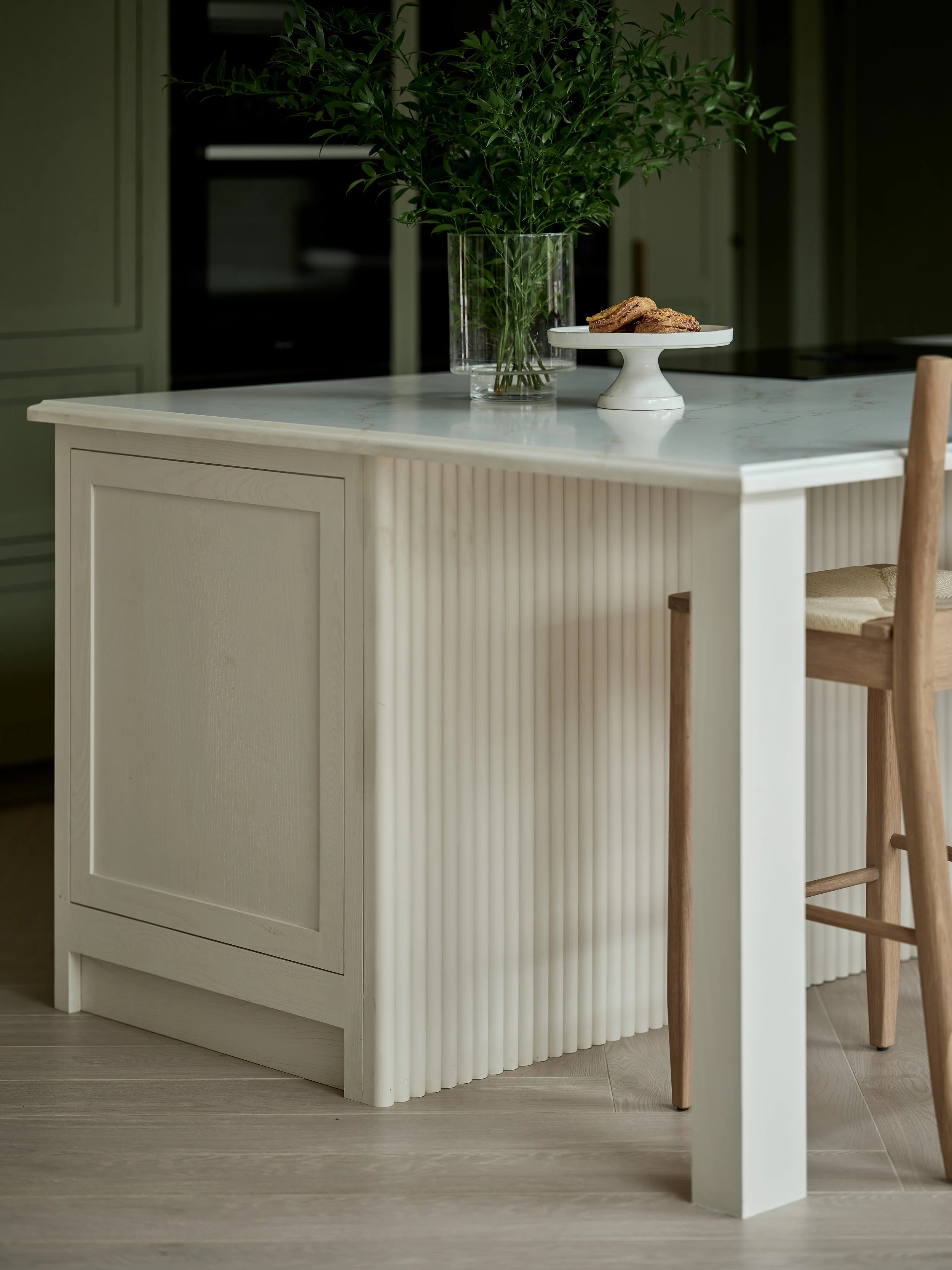 A white kitchen island with a marble top featuring a large green leafy plant in a glass vase and a white cake stand with baked cookies on top. Part of a wooden chair is visible in the foreground on the right.
