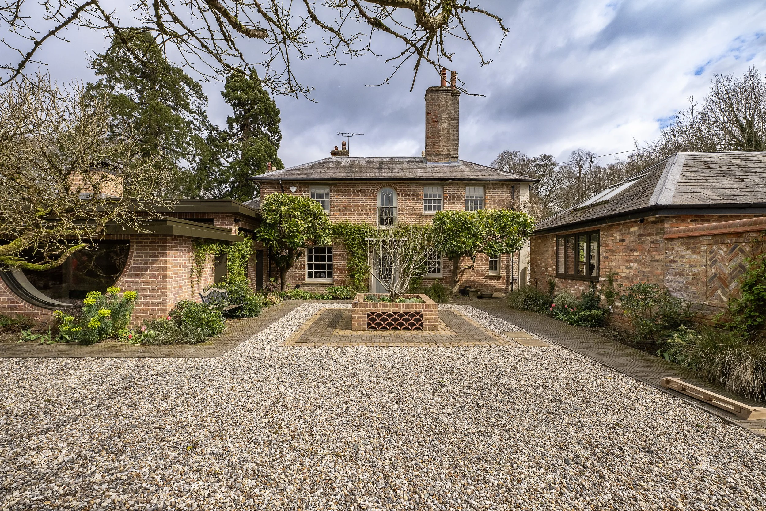 Brick house with smokestack, surrounded by garden with trees and plants, gravel ground, cloudy sky.