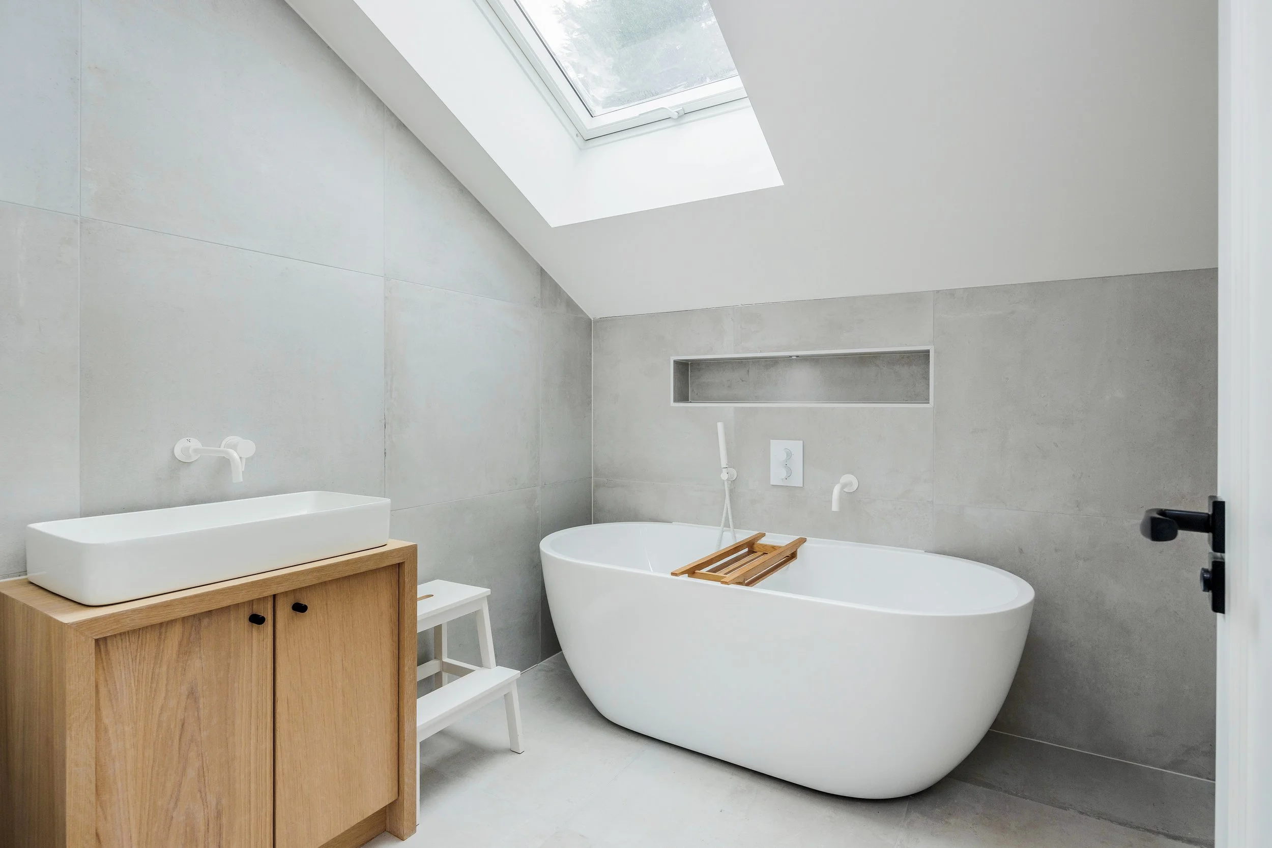 A modern bathroom with a sloped ceiling and skylight window, featuring a white freestanding bathtub with a wooden bath tray, a wooden vanity with a white rectangular sink, and minimal decor.