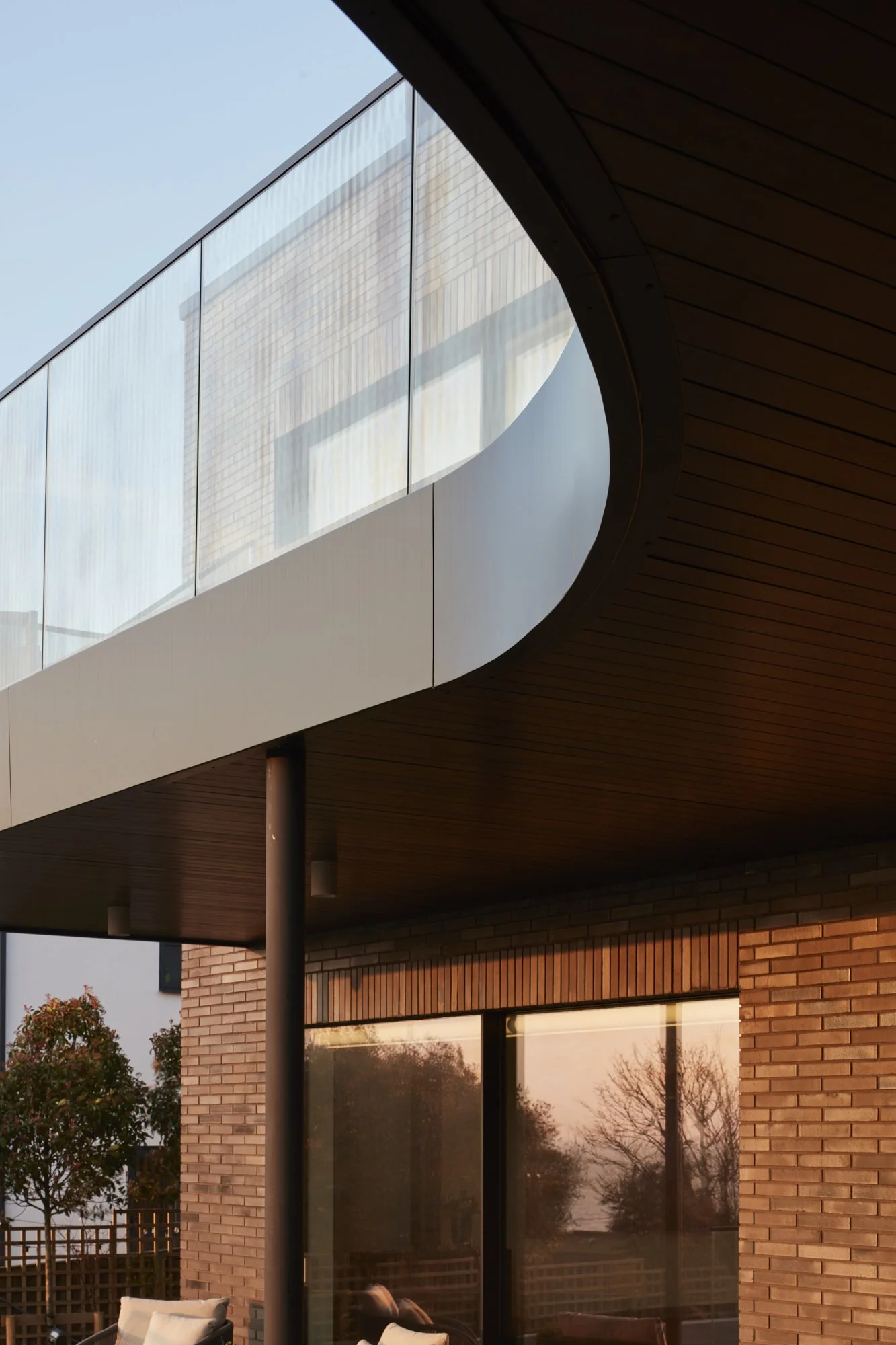 Exterior of a modern house with large glass window, brick wall, and wooden ceiling, during sunset with trees and outdoor furniture visible.