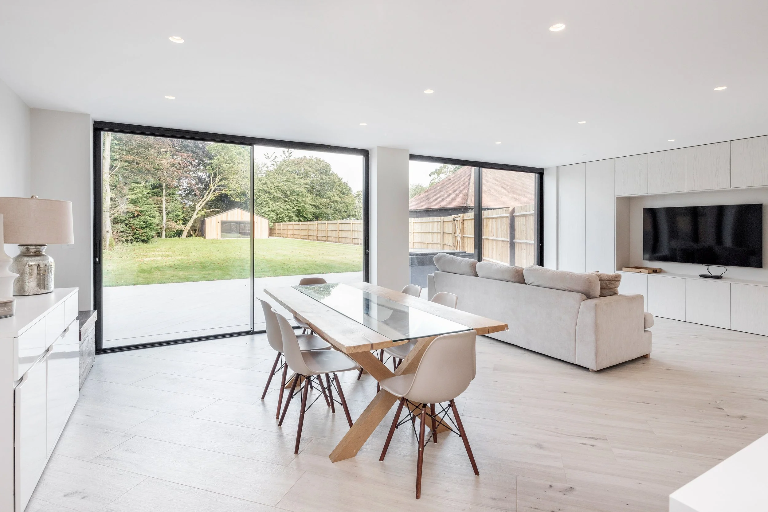 Modern living room with large sliding glass doors opening to a backyard with a wooden shed and trees, includes a dining table with chairs and a beige sofa facing a wall-mounted TV.