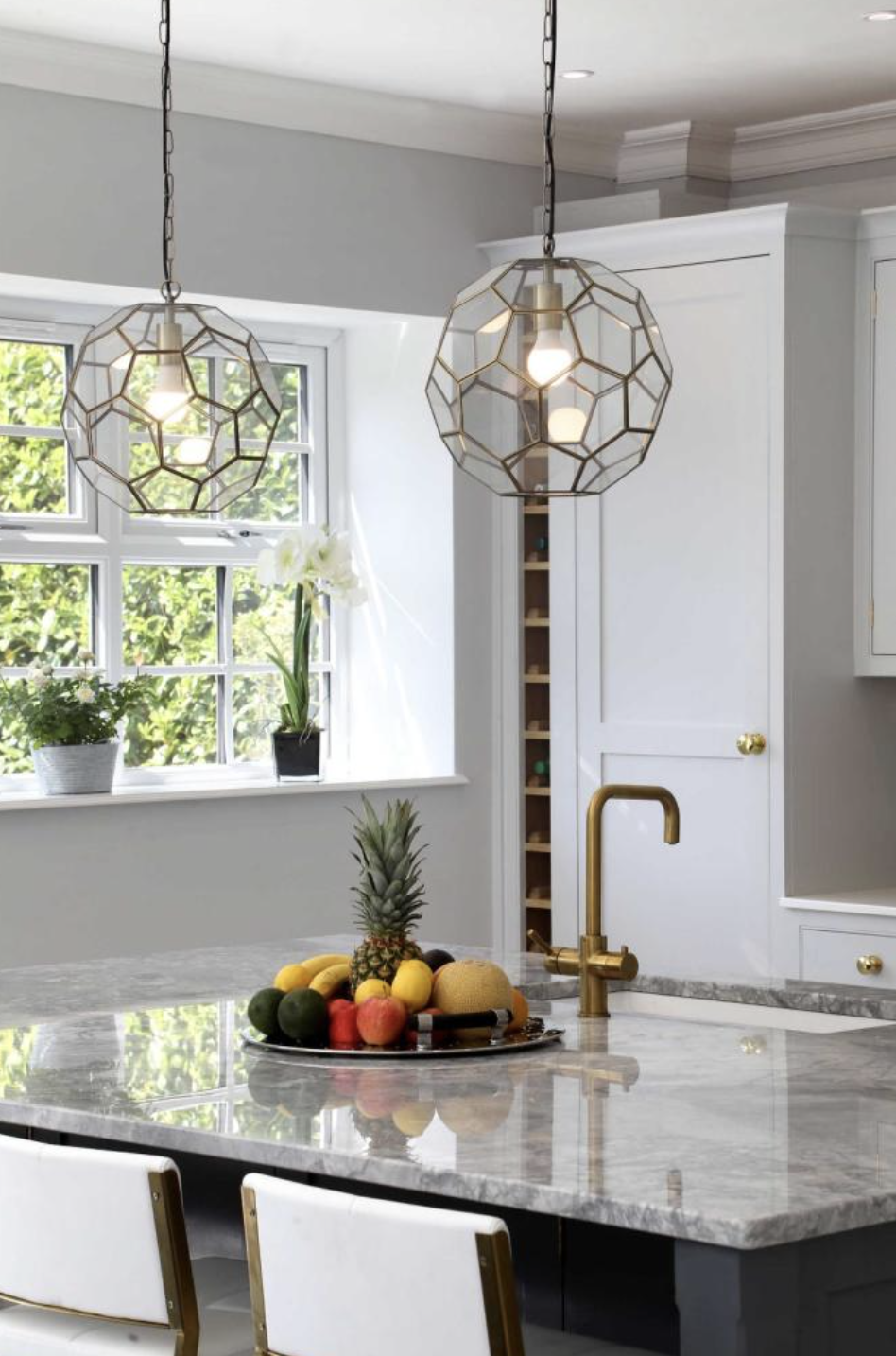 A modern kitchen with two geometric pendant lights hanging over a marble countertop, a window with greenery outside, and a bowl of tropical fruits including pineapple, bananas, apples, and melons on the counter.