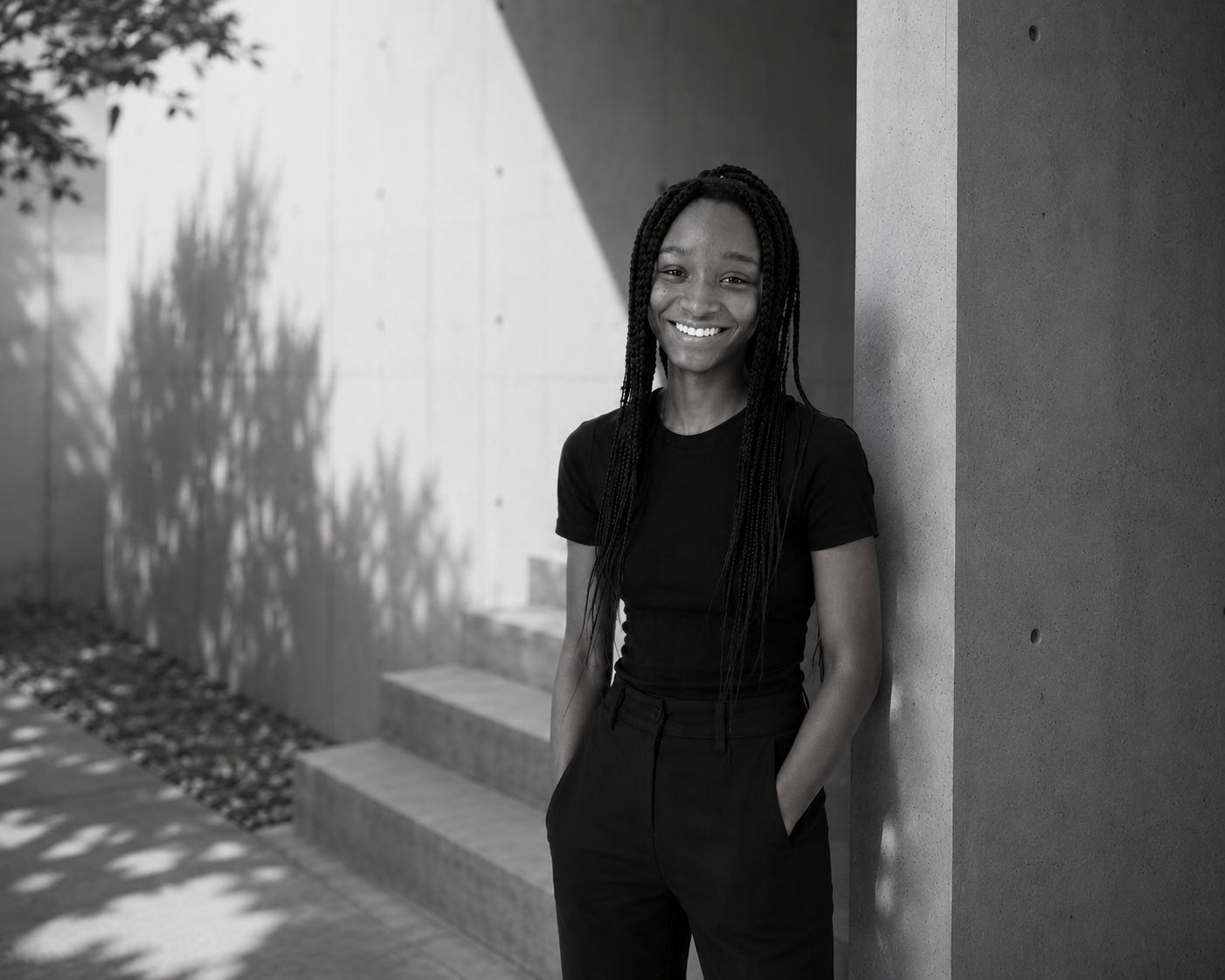 A young woman with long braids, smiling, standing outdoors against a modern concrete wall, wearing a black t-shirt and black pants.