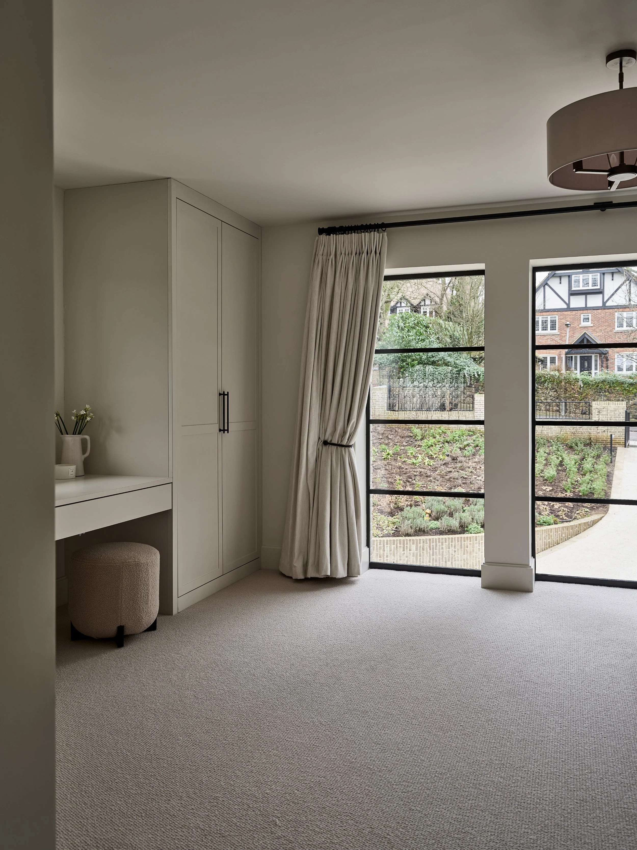 Interior view of a bright room with beige carpet, white walls, a built-in wardrobe, a small vanity desk with an upholstered stool, and large windows with cream curtains overlooking a garden and neighboring houses.