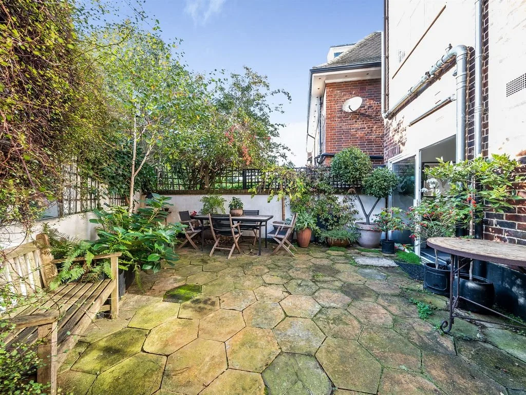 A small outdoor patio with hexagonal stone paving, surrounded by green plants and shrubs. There is a wooden bench on the left and a round table with several chairs in the center. Potted plants and a lattice fence are along the back, and part of a bri