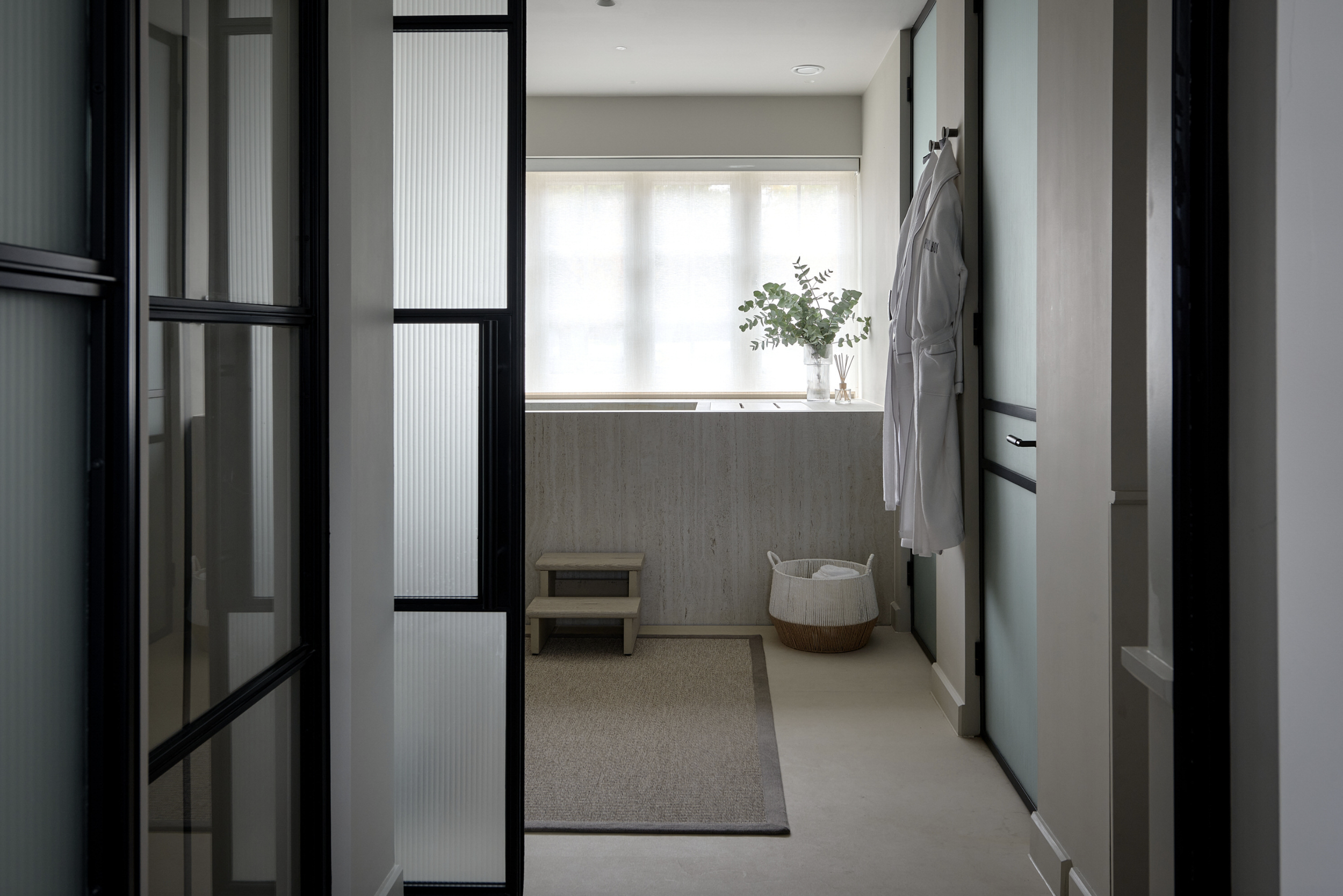 A view inside a modern bathroom with a bathtub, a small stool, a basket, a white robe hanging, and a potted plant near a large window with sheer curtains.