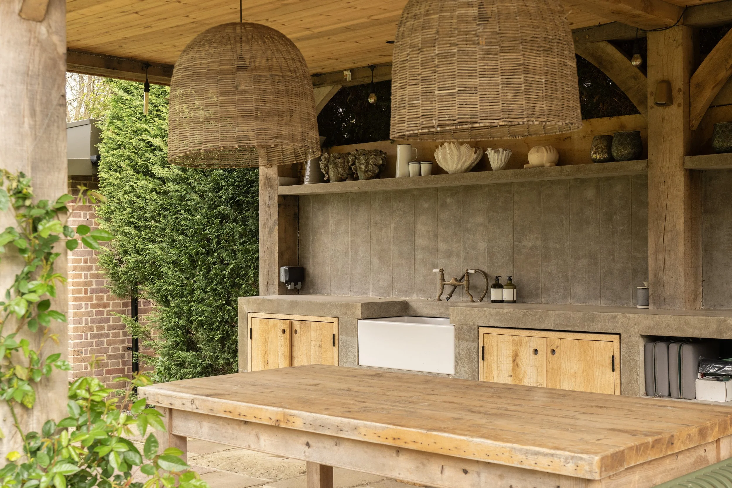 Outdoor kitchen area with a concrete countertop, wooden cabinetry, and shelves with vases and decorative items. Hanging wicker pendant lights illuminate the space, surrounded by greenery and brick walls.
