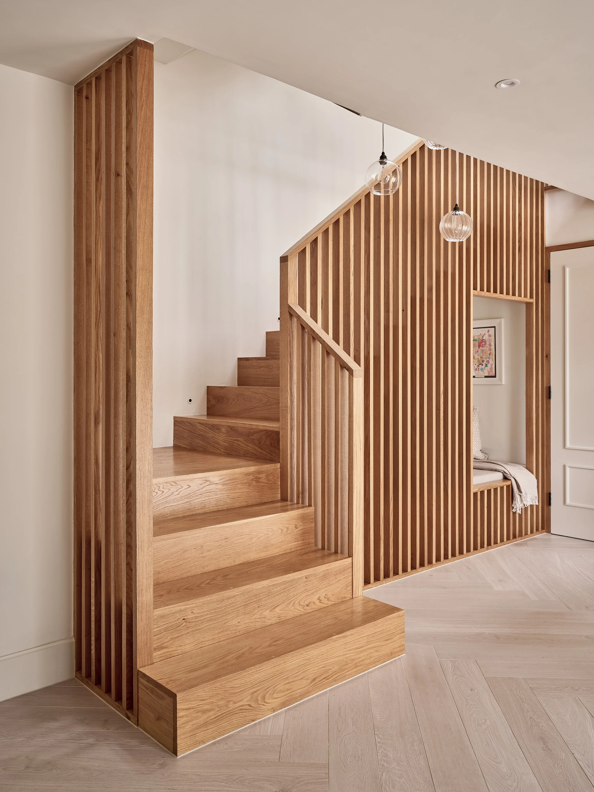 Interior view of a wooden staircase and a wooden screen with vertical slats, near a small seating nook with a framed artwork on the wall.