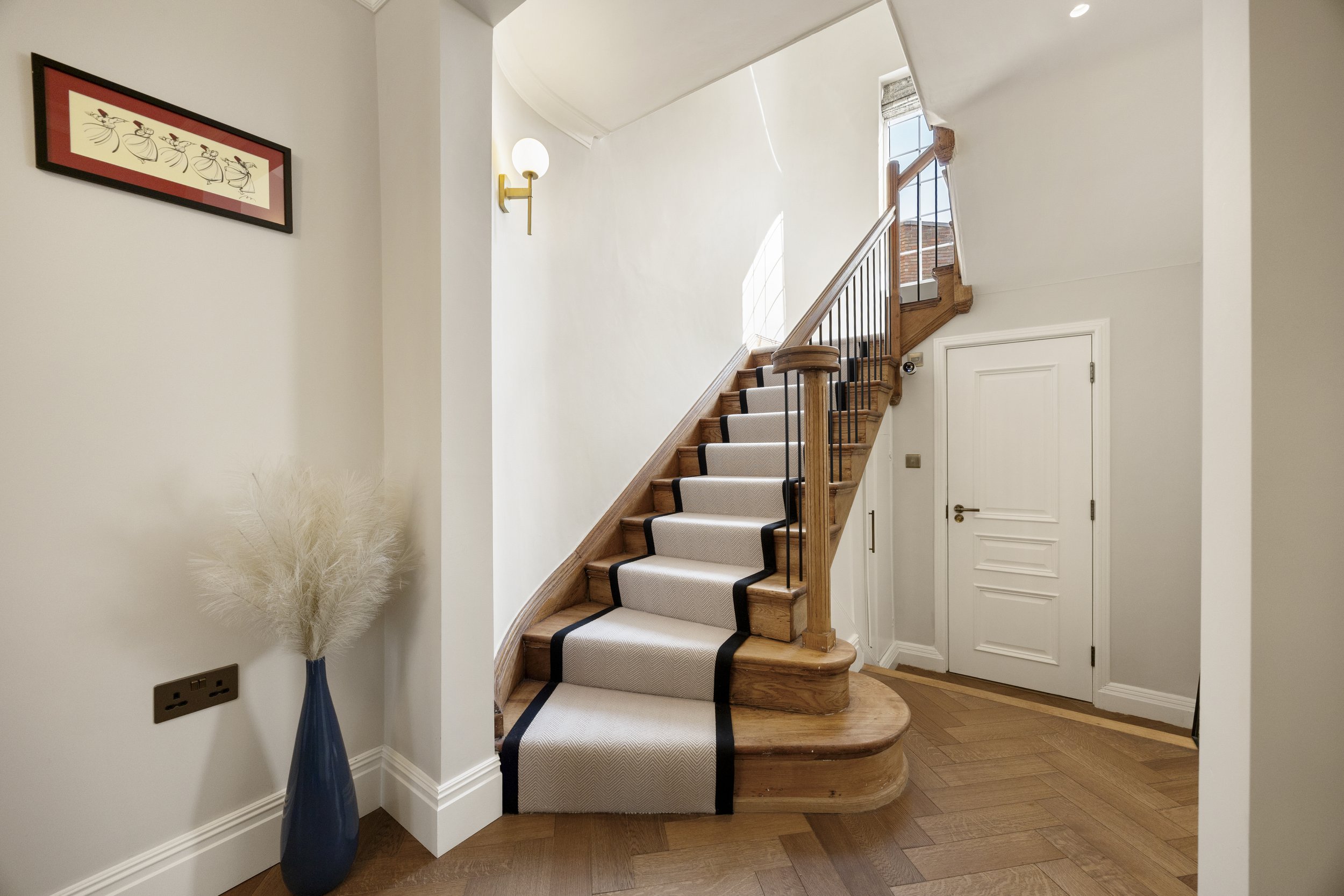 A wooden staircase with a beige runner and black trim, a blue vase with white pampas grass beside a white wall, and a framed artwork featuring stylized dancers.
