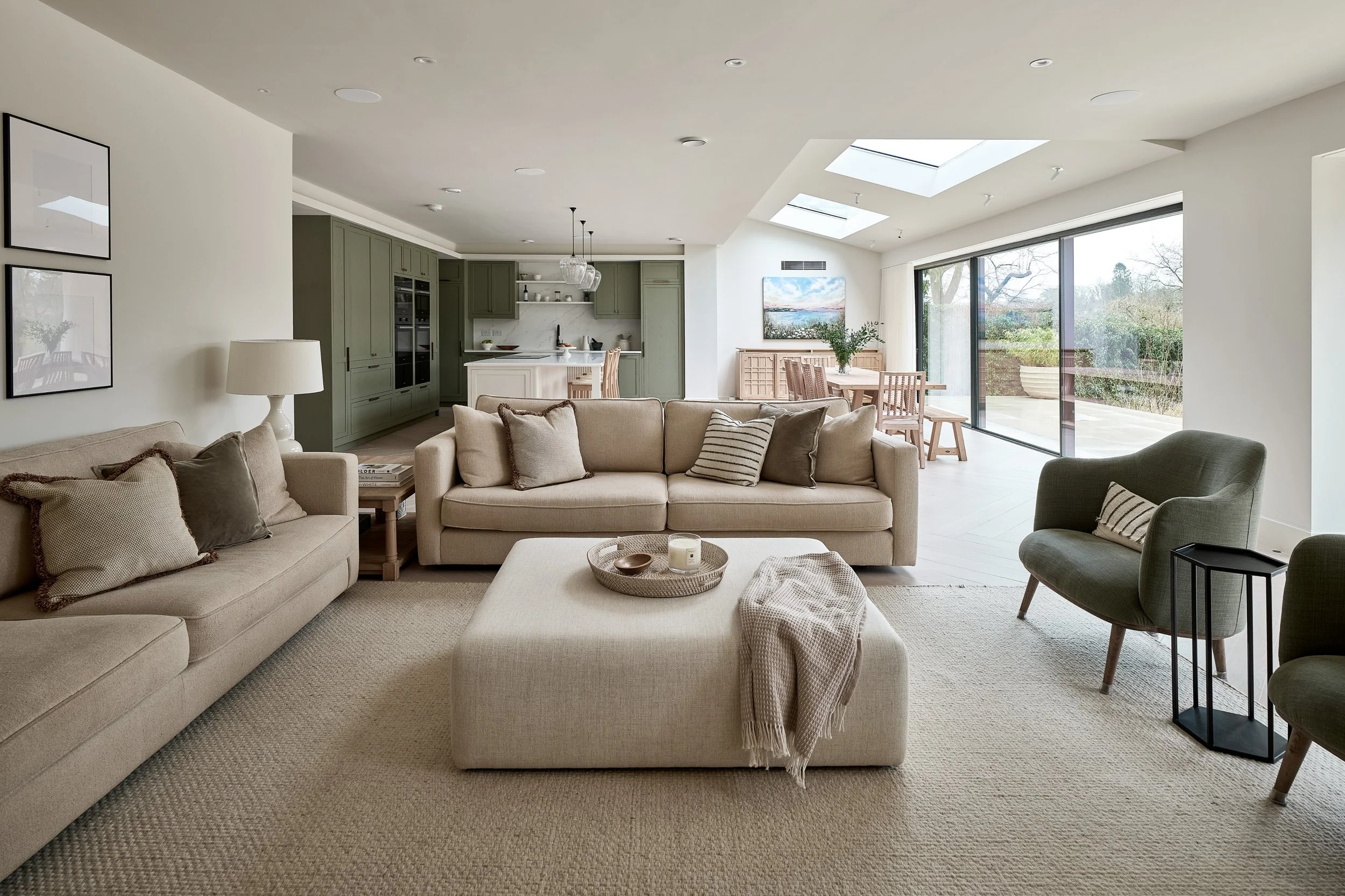 A spacious, modern living room with beige sofas, a stone coffee table, and light-colored carpet, open to a dining area and a kitchen with green cabinets.
