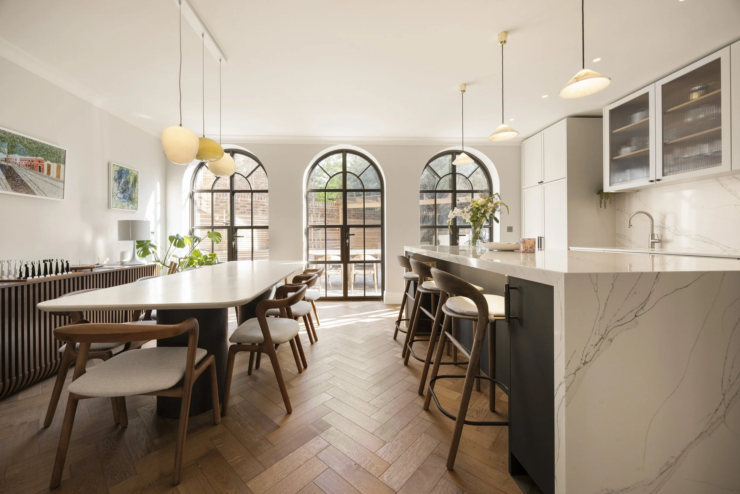 A modern kitchen and dining area with arched windows, a large marble island, wooden chairs, pendant lights, and artwork on the wall.