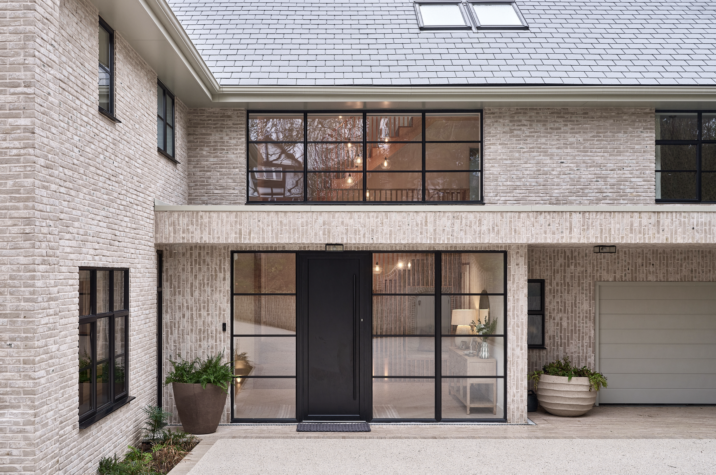 Front view of a modern brick house with large glass doors and windows, potted plants, and a paved patio area.