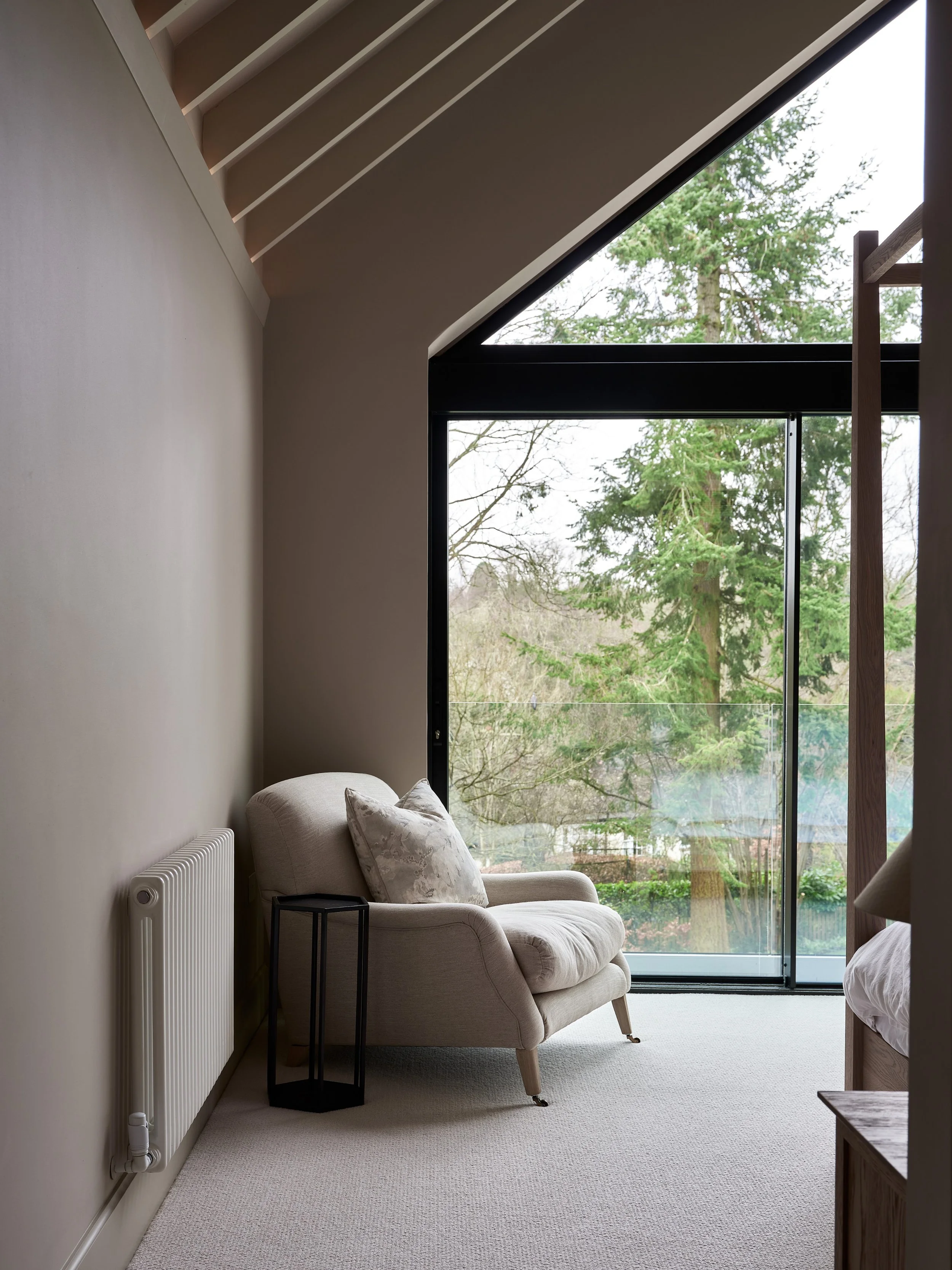 A cozy living room with a beige armchair, pillow, and side table near a large window overlooking a green outdoor landscape with trees.