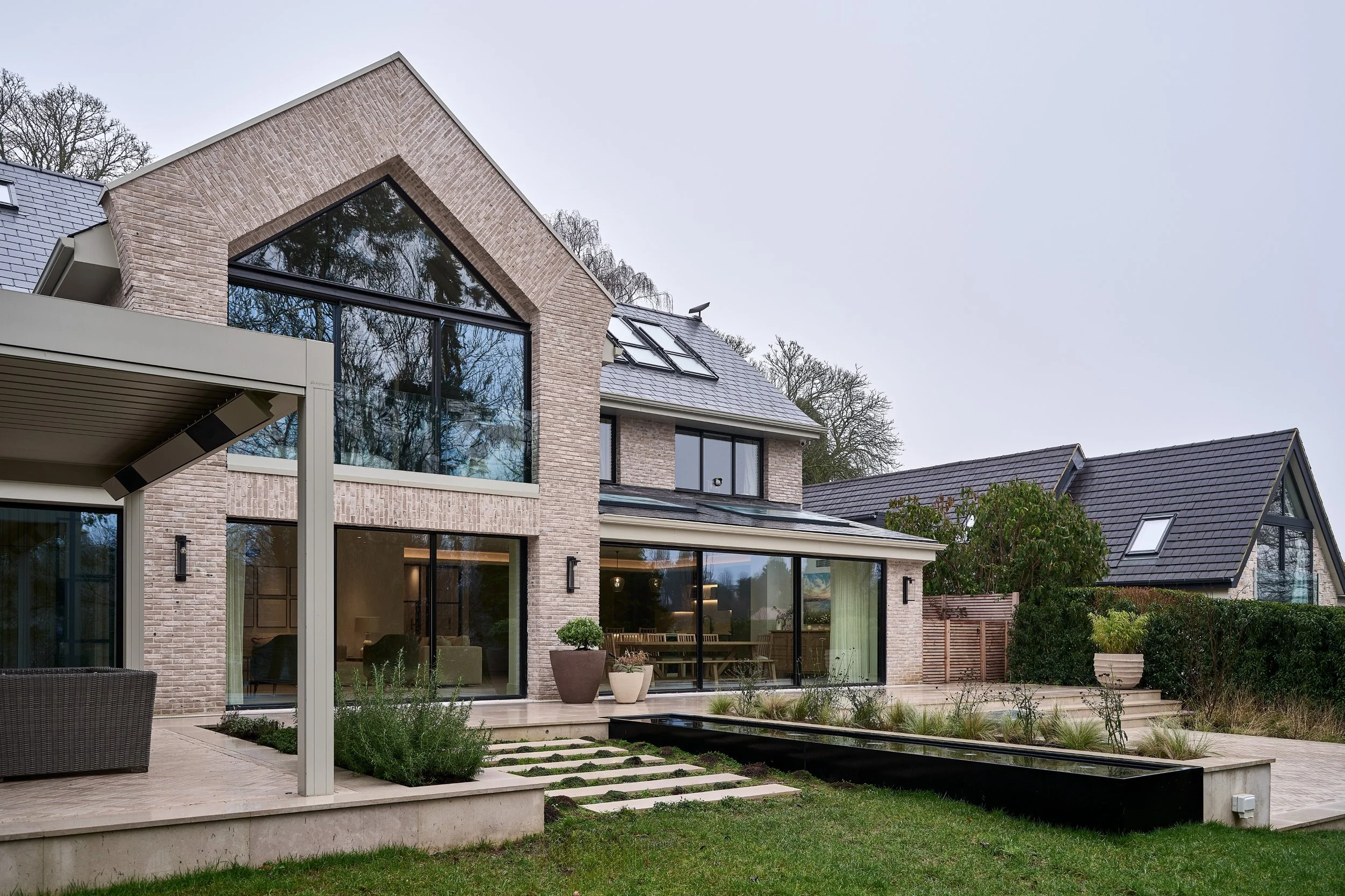 Modern house with large glass windows, brick exterior, and a sloped roof, with a manicured backyard featuring potted plants, a patio, and a small pond.