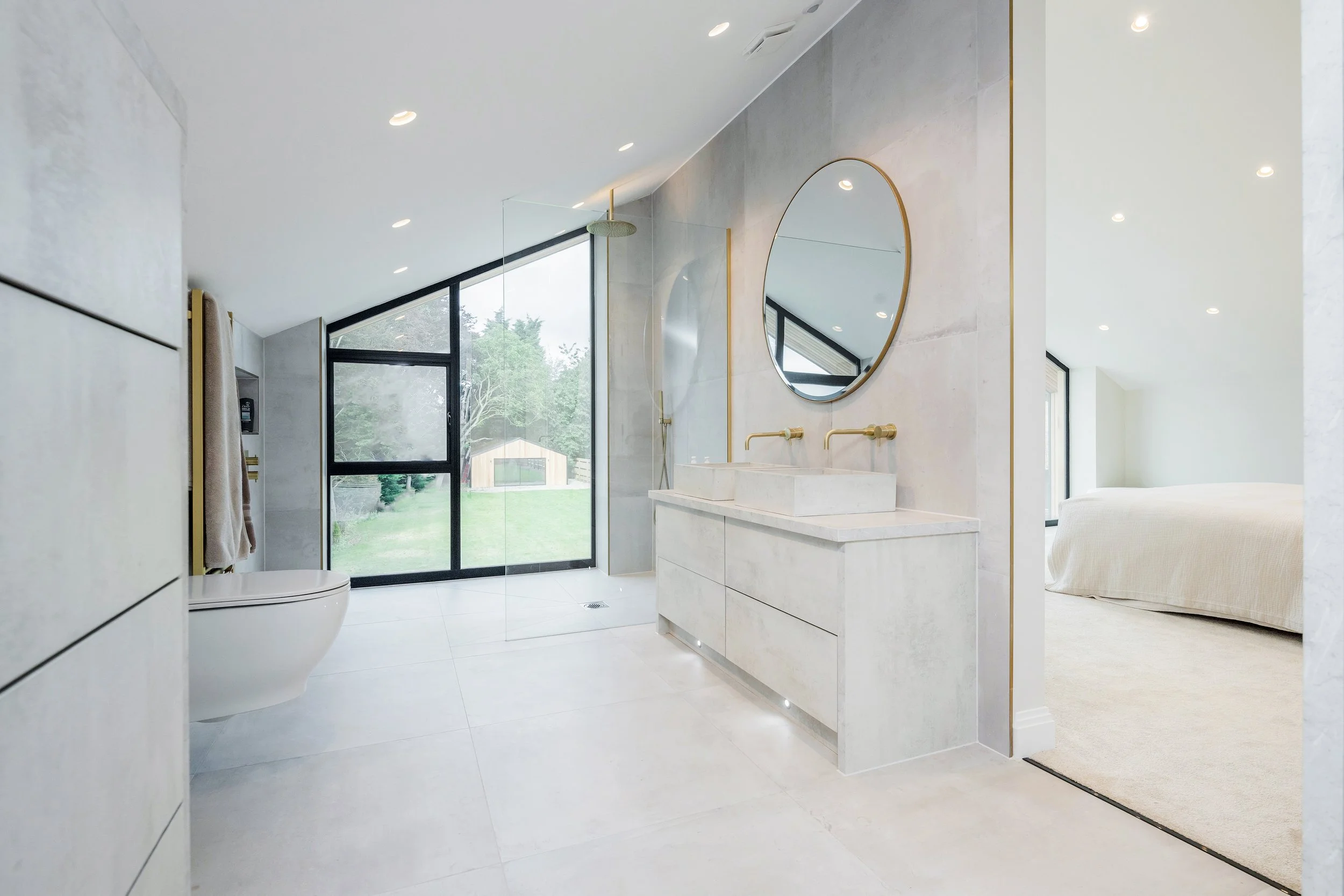 Modern bathroom with large window, round mirror, double sink vanity, walk-in shower, and sliding door to outdoor view.