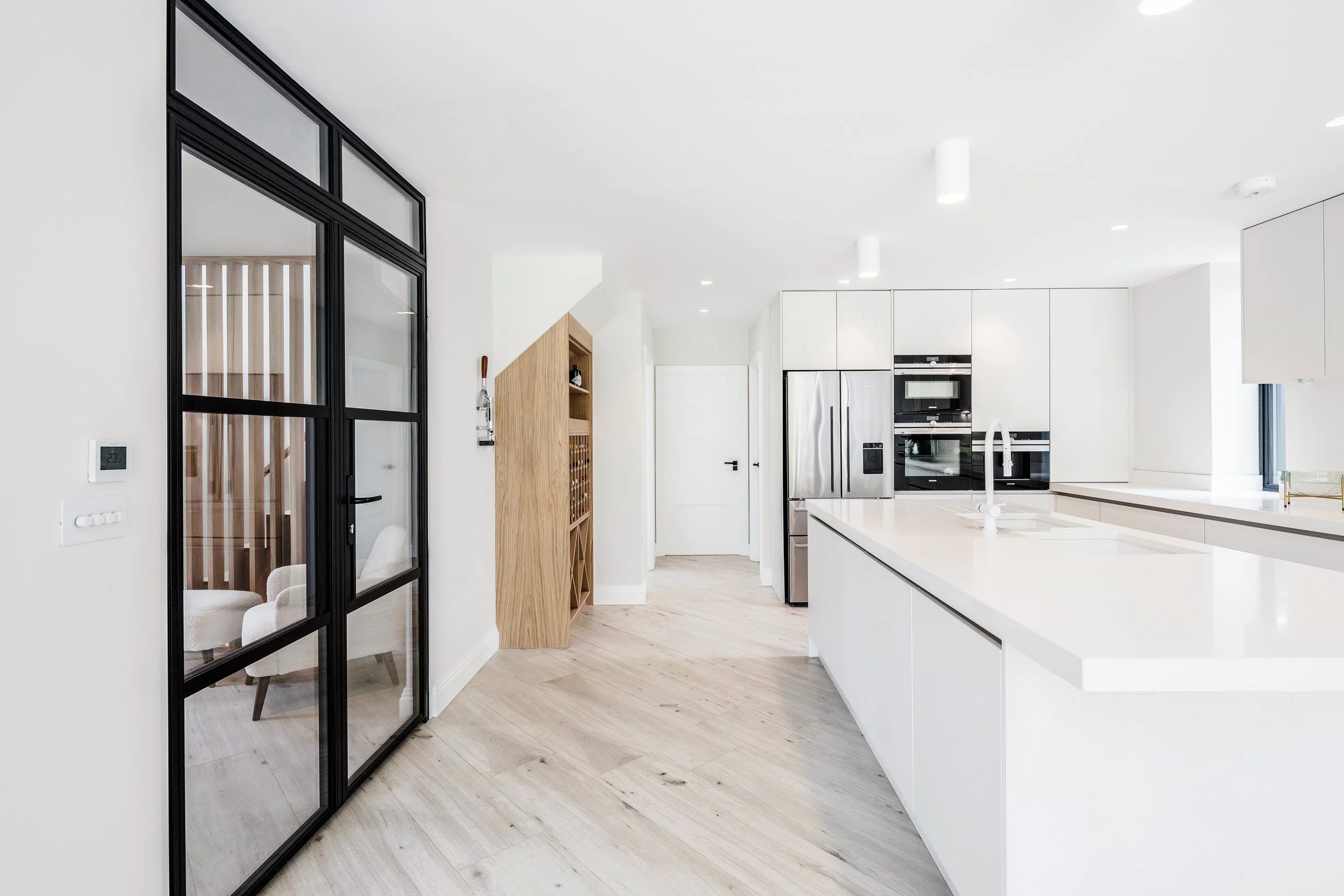 Modern kitchen with white cabinets, black-framed glass door, stainless steel refrigerator, and integrated oven, with light wood flooring and minimal decor.