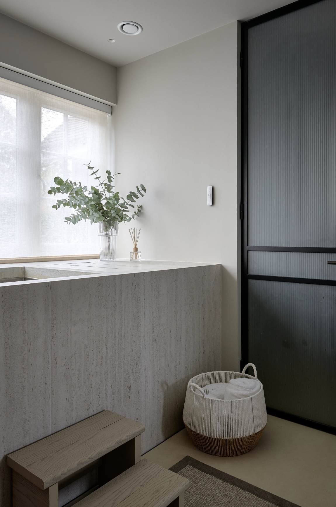 Minimalist kitchen with a white countertop, a glass vase with greenery, a diffuser, and a window with sheer curtains, next to a black sliding door and a white basket with towels.