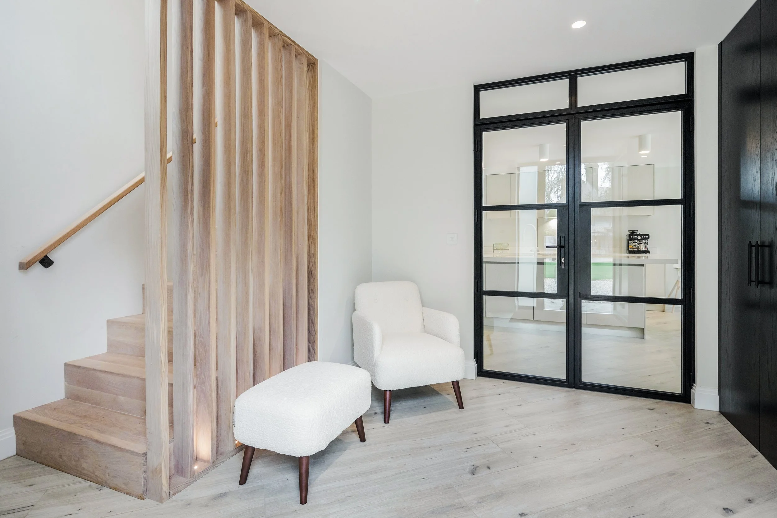 A modern interior space with a staircase on the left made of light wood and a black-framed glass door on the right. There are two white cushioned chairs with wooden legs near the door, and part of a kitchen or living area is visible through the glass