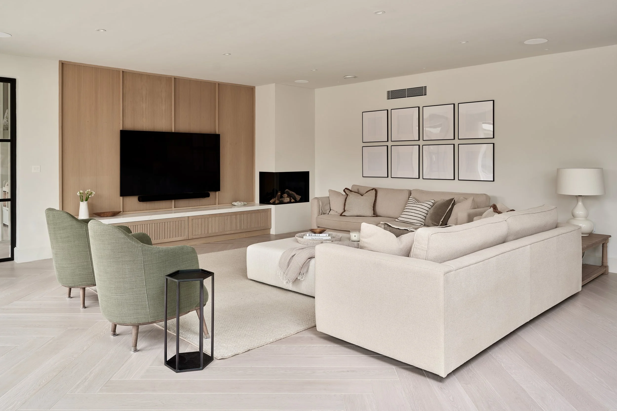 Modern living room with beige sofa, green armchairs, black side table, light wood flooring, wall-mounted TV, white walls, a picture frame collage, and a fireplace.