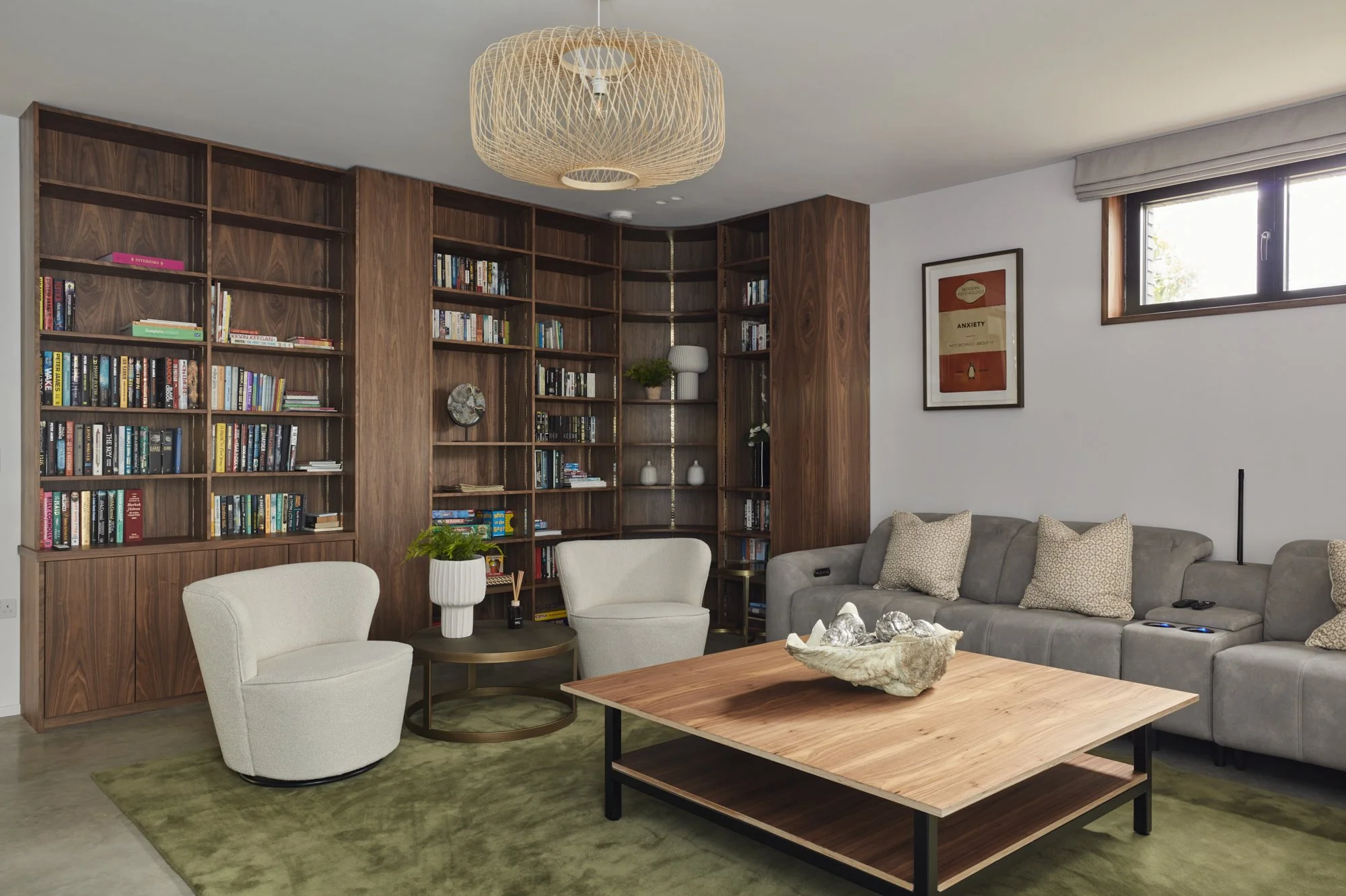 A cozy living room featuring a large wooden bookshelf filled with books, a gray sofa with patterned throw pillows, a round white armchair, a patio table with a potted plant, a rectangular wooden coffee table with decorative items, a wall with a frame