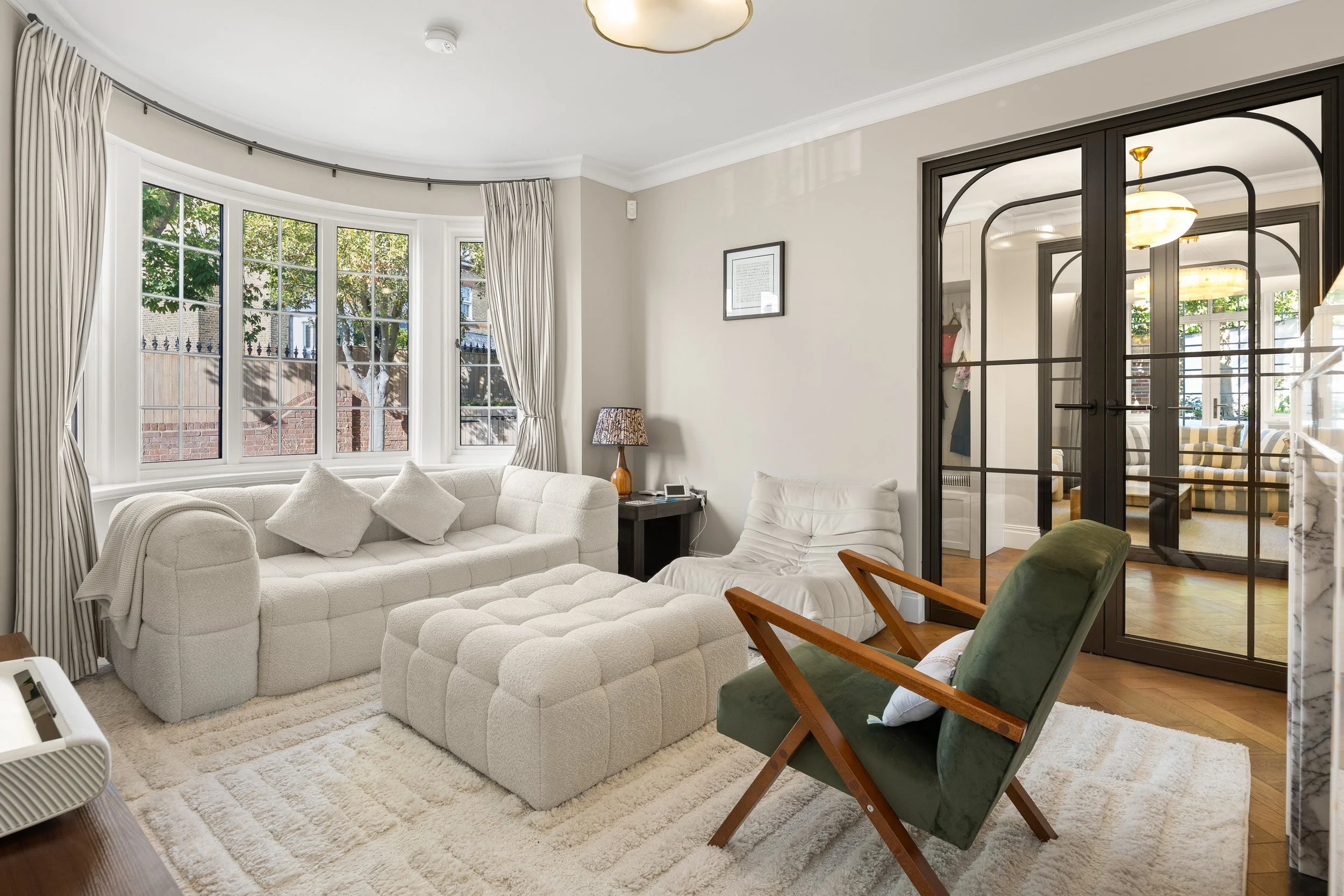 Living room with large bay window, white sofa with multiple cushions, green armchair, end table with lamp, and interior double doors leading to adjacent room with striped cushions and other furnishings.