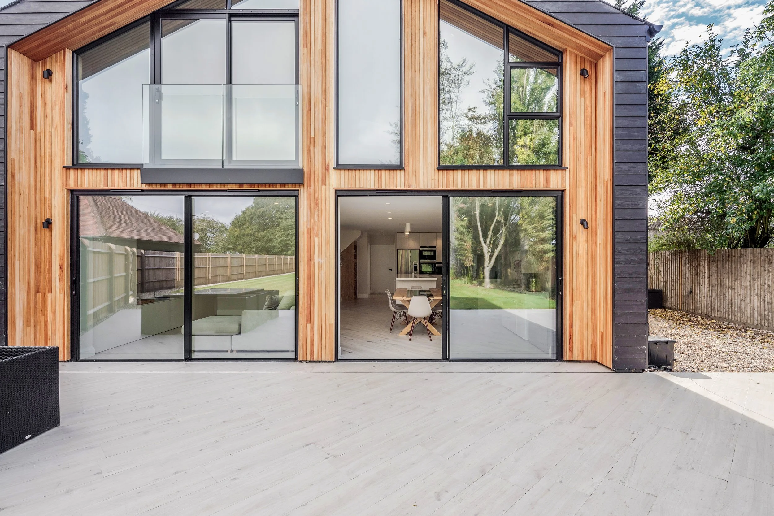 A modern multi-story house with large windows and sliding glass doors, featuring a wooden exterior and a spacious outdoor patio with a light-colored wooden floor.