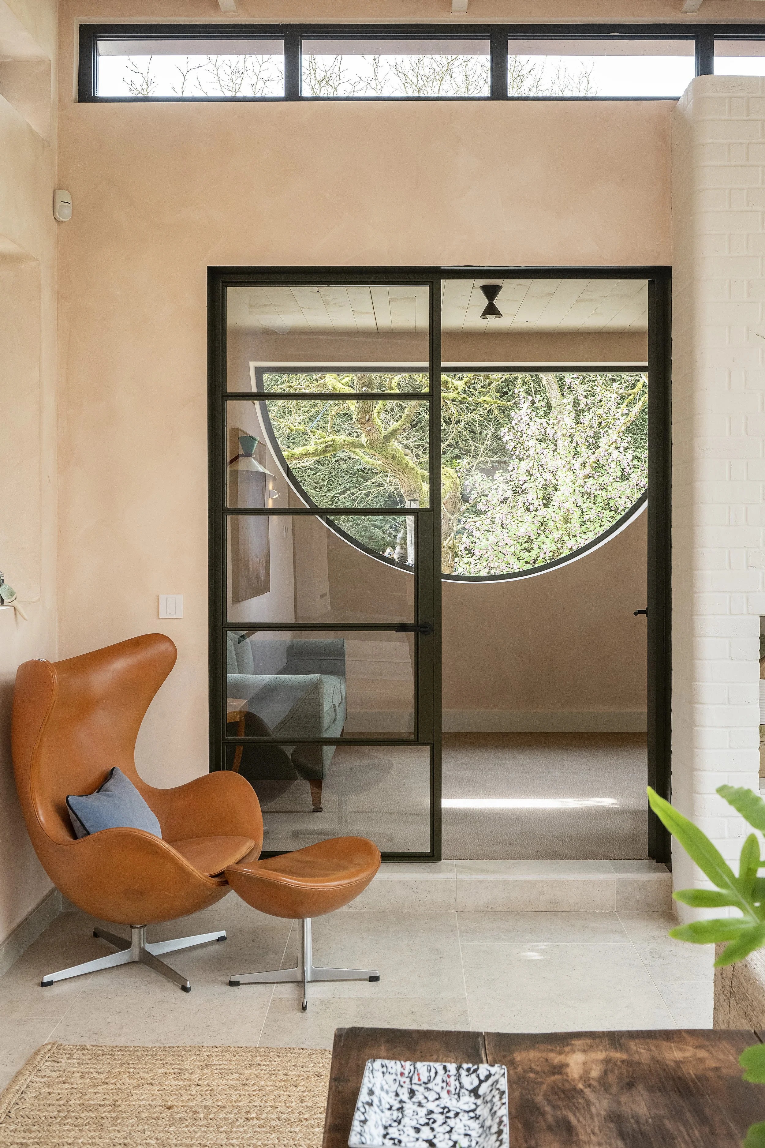Modern living space featuring a black-framed glass door, a leather lounge chair with a matching footrest, a small gray sofa, and a view of a garden with a flowering tree through the window.