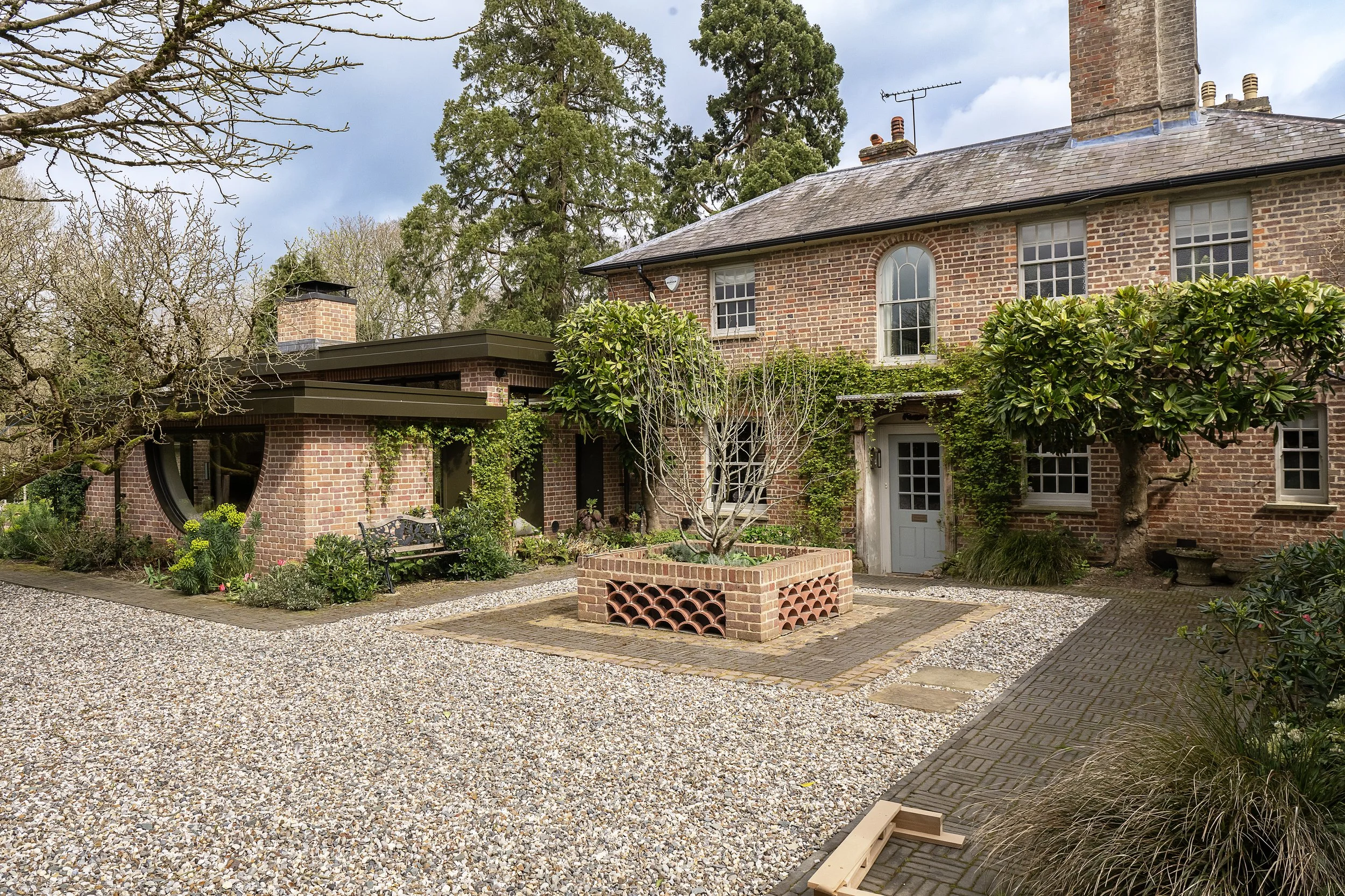 A brick house with multiple windows, surrounded by trees and a garden. There is a gravel pathway in the foreground and a brick plant bed with a small tree. The sky is partly cloudy.