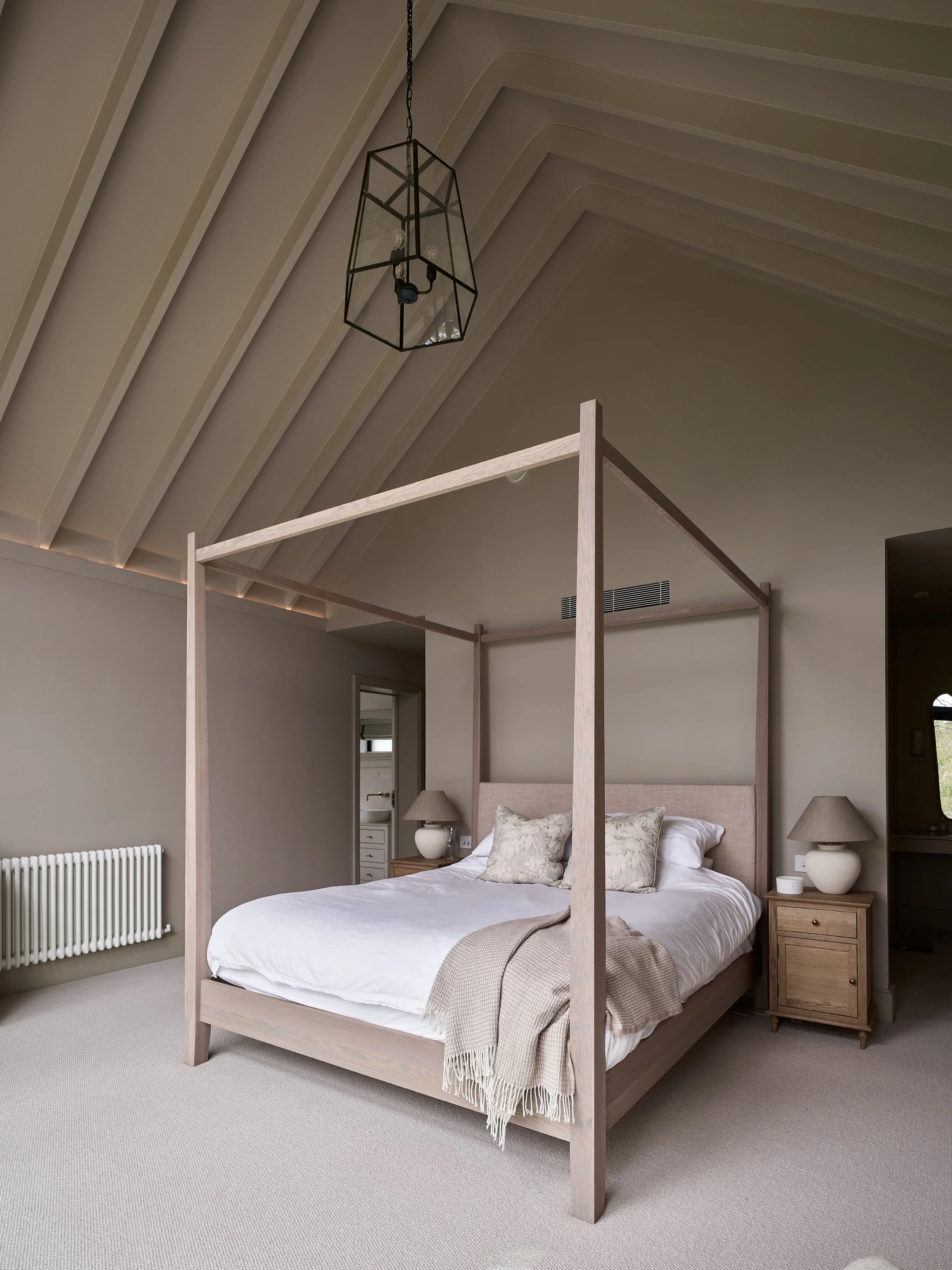 A bedroom with a large bed, bedside tables, lamps, and a bed frame with four posts. The room has vaulted ceiling with a hanging light fixture, grey walls, a radiator, and a doorway to a bathroom.