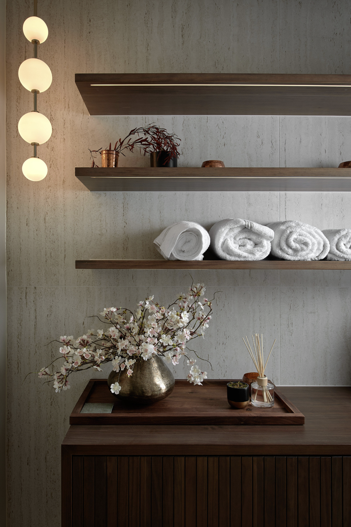 A bathroom or spa area with three floating wooden shelves mounted on a textured beige wall. The shelves hold rolled white towels, small copper and black pots, and decorative items. To the left, a vertical wall sconce with four evenly spaced round whi