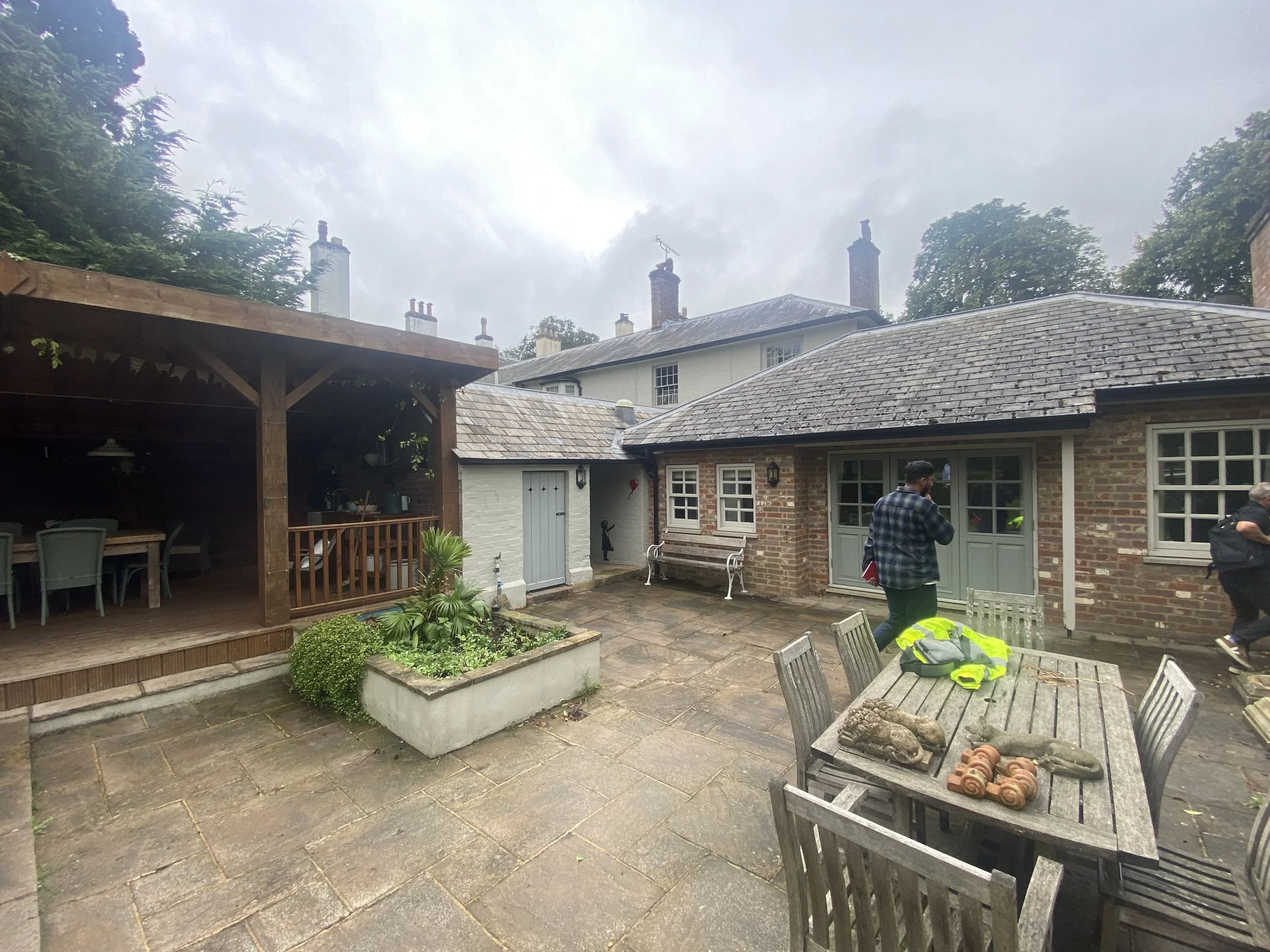 A backyard patio area with outdoor dining furniture, a wooden pergola, and a brick house with a tiled roof. There are people standing and walking, and a table with decorative objects. Overcast sky and neighboring houses are visible.