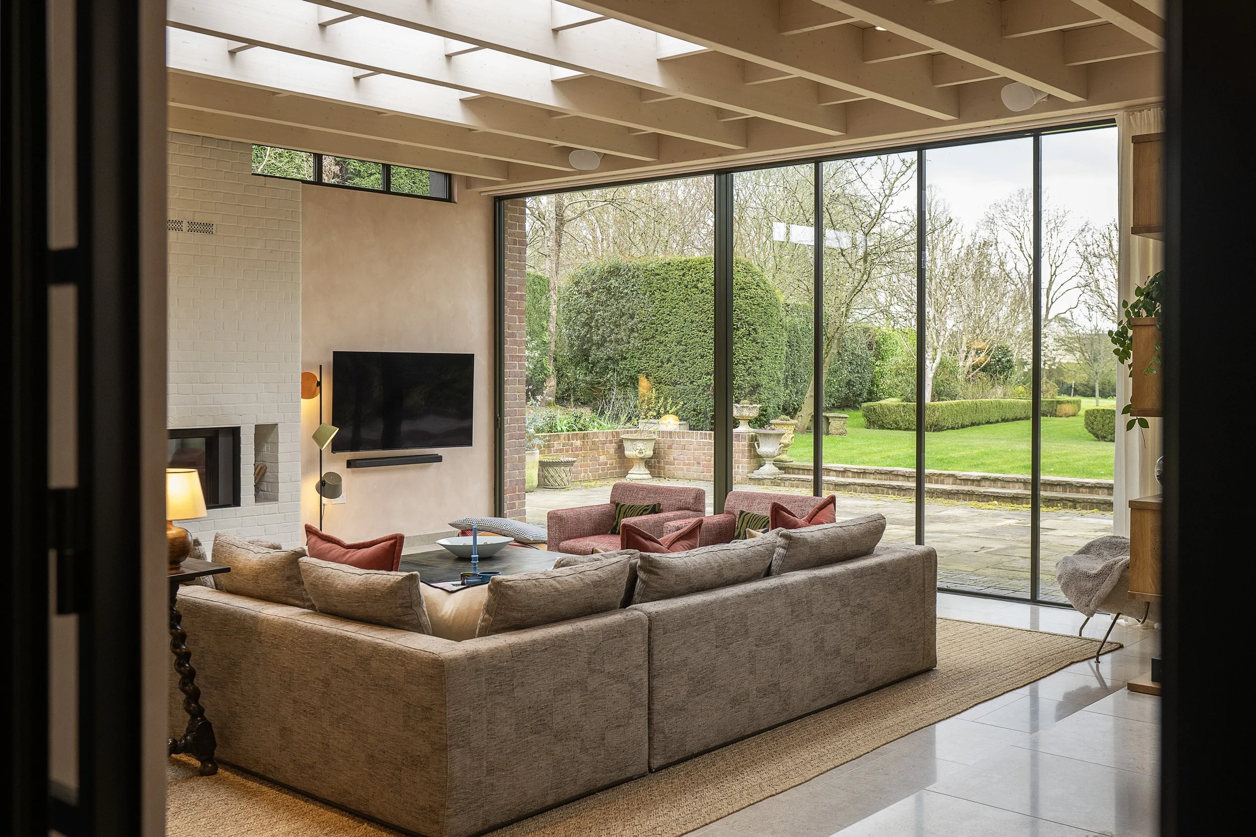 A cozy living room with large floor-to-ceiling windows showing an outdoor garden with trees, bushes, and decorative planters. The room has a beige sofa with red and green pillows, a coffee table with a bowl and a candlestick, and a fireplace with a T