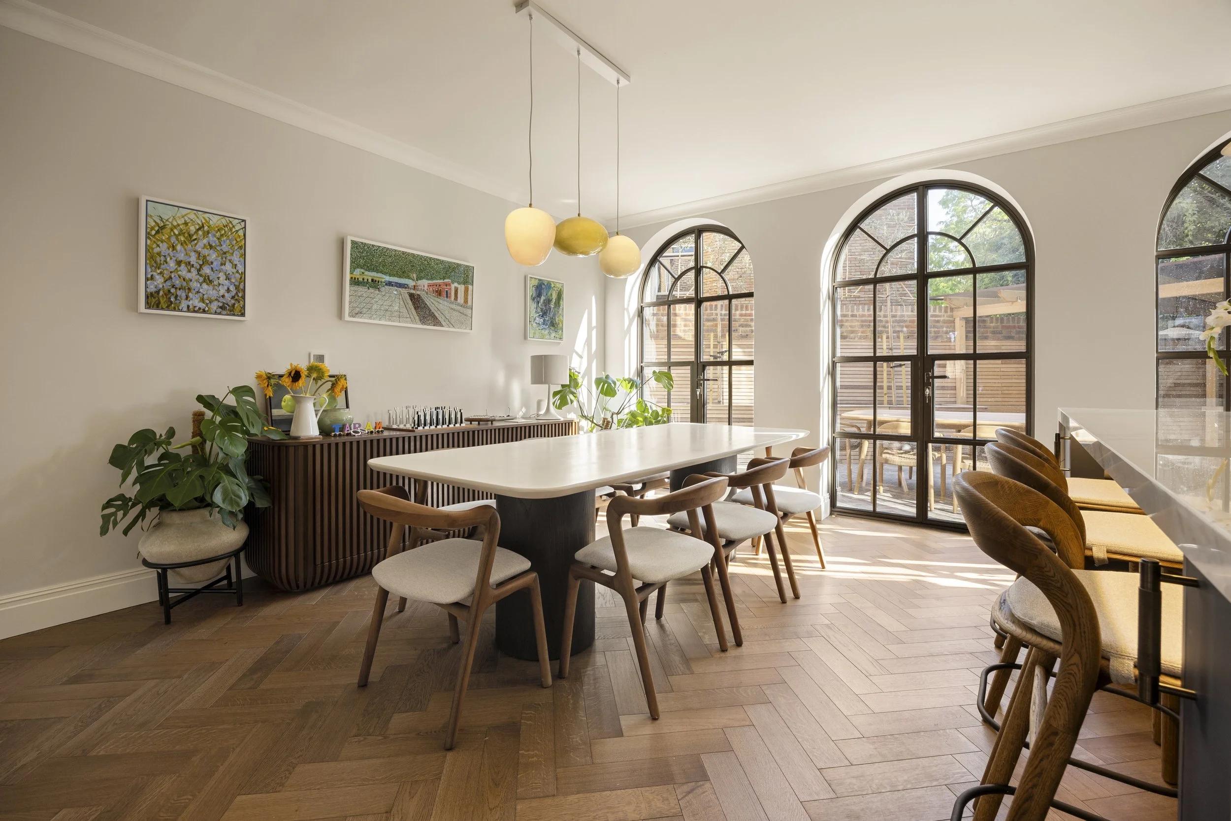 Bright dining room with large arched windows, wooden floor, white walls, modern art, circular chandelier, plants, and wooden chairs around a white table.