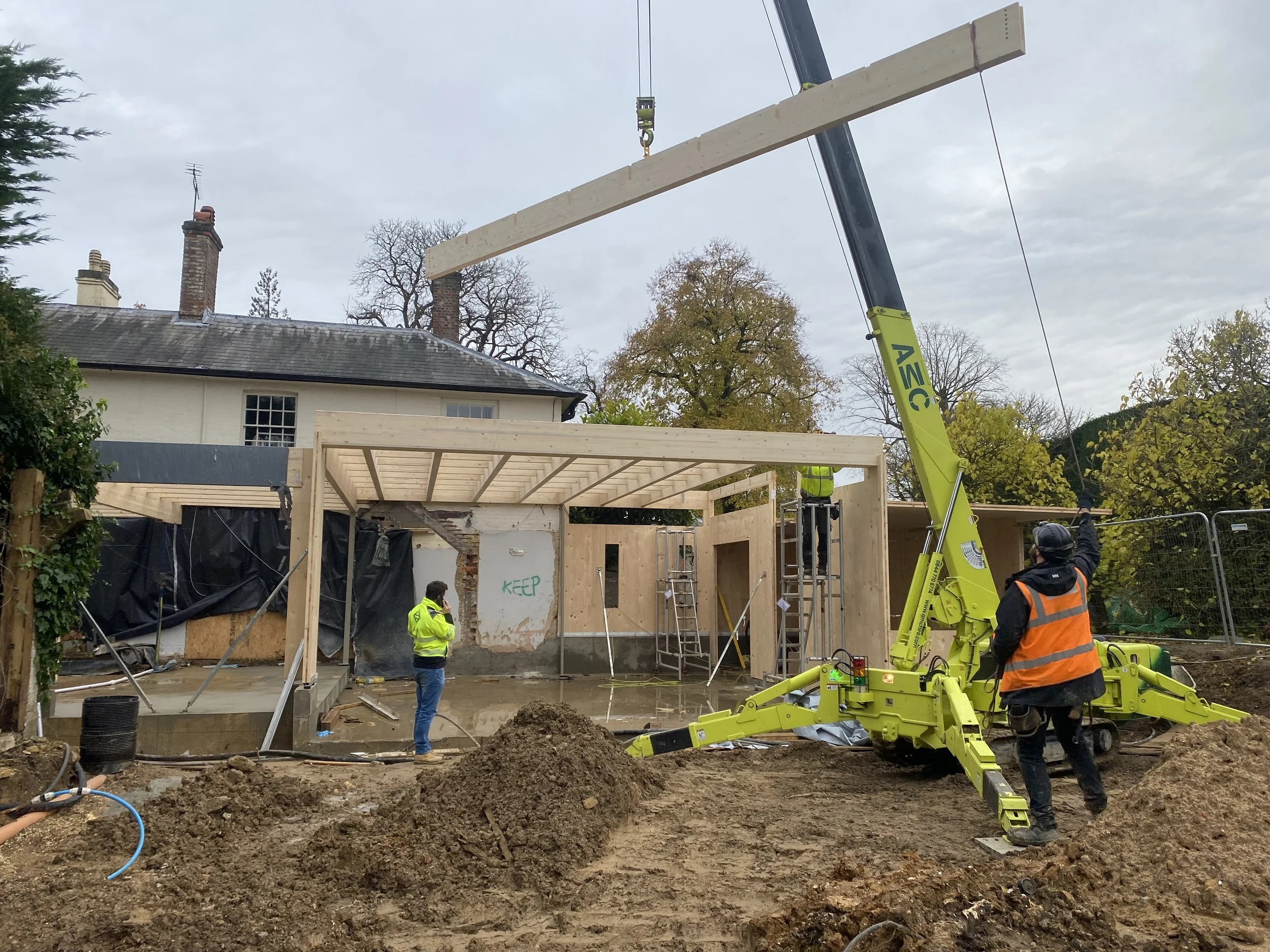 Construction workers at a building site with a crane lifting a large wooden beam, working on framing and foundation for a new structure.