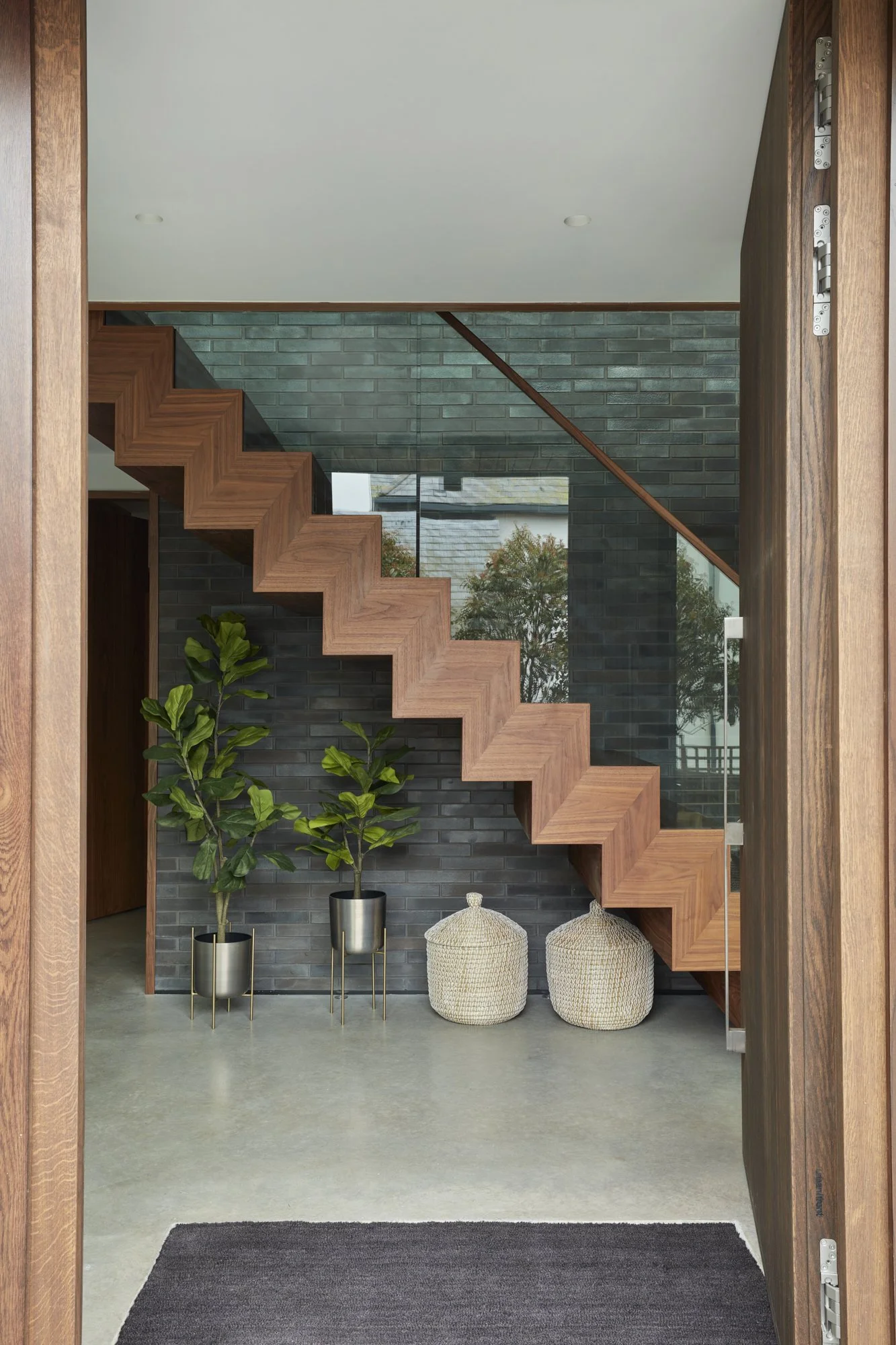 View of modern interior with wooden staircase, two large potted plants, woven baskets, and gray brick wall. Glass window shows outside trees.