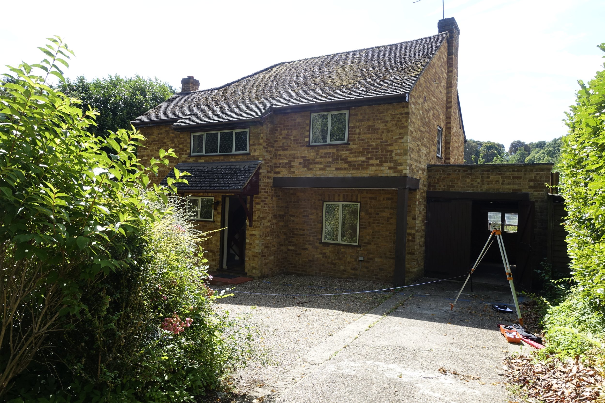 A brick house with a sloped roof, surrounded by green bushes, with tools and equipment on the driveway, under a partly cloudy sky.