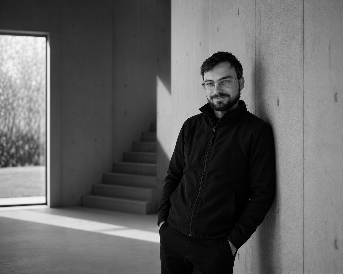 A man with glasses and a beard leaning against a concrete wall inside a modern building, with stairs and a window showing trees outside in the background.