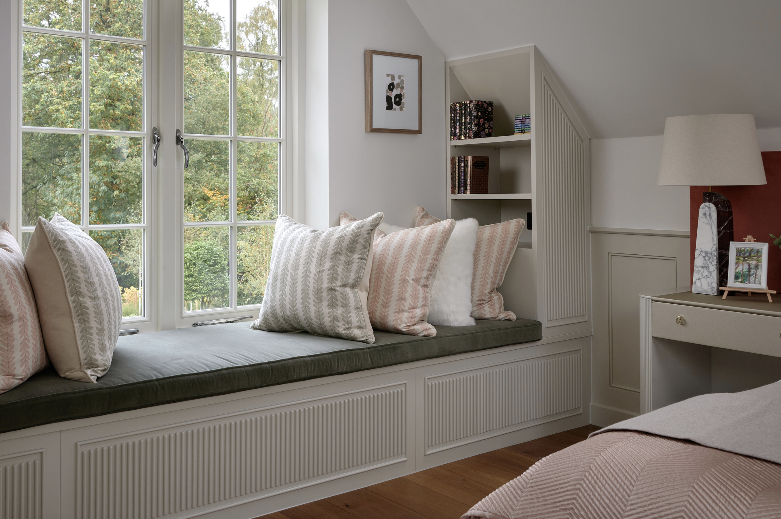 A window seat with pillows in a cozy bedroom with large windows showing trees outside, a built-in shelf with books, and a bedside table with a lamp and photo frame.