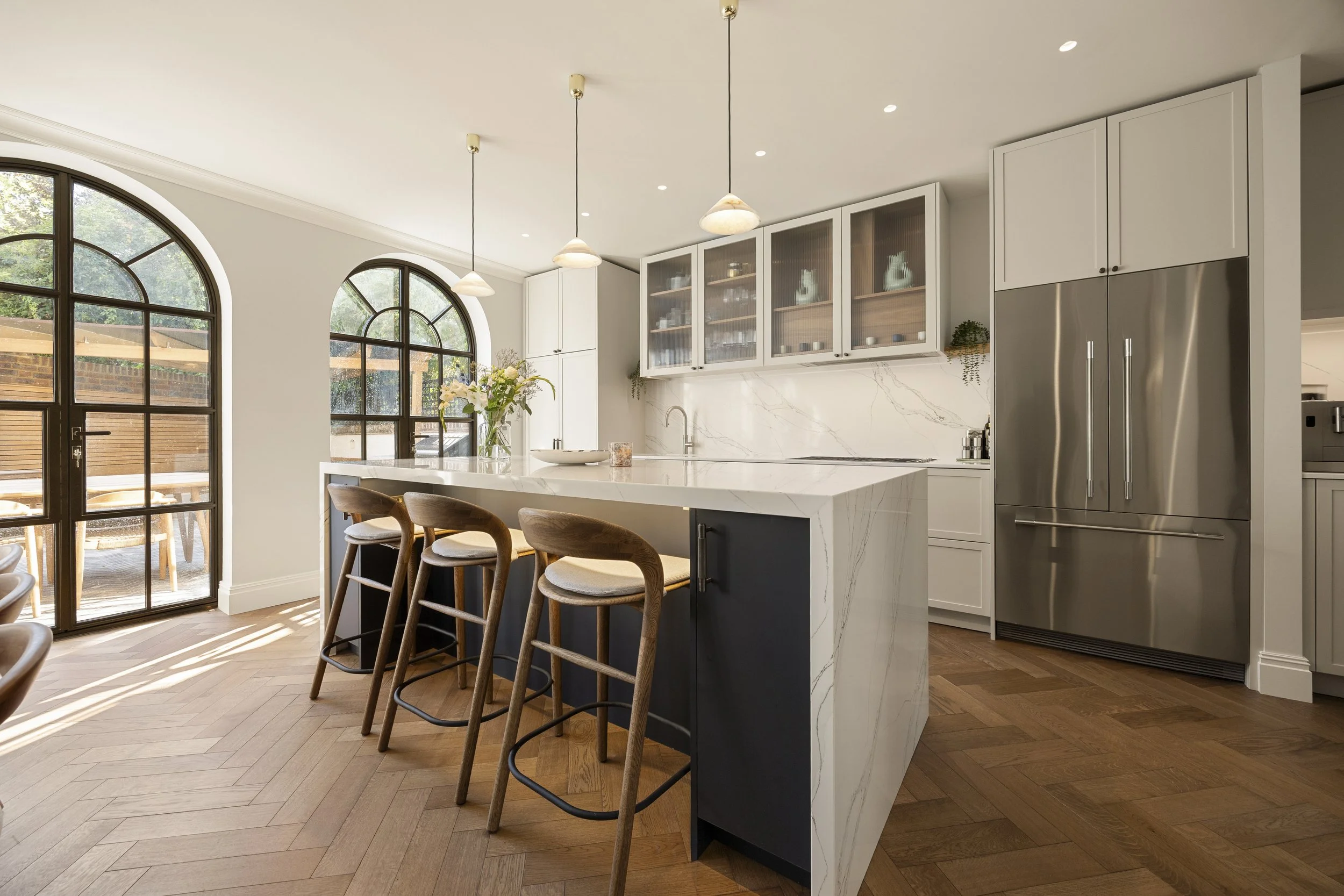 Modern kitchen with white cabinetry, marble island, wooden barstools, black accents, large arched windows, and hanging pendant lights.
