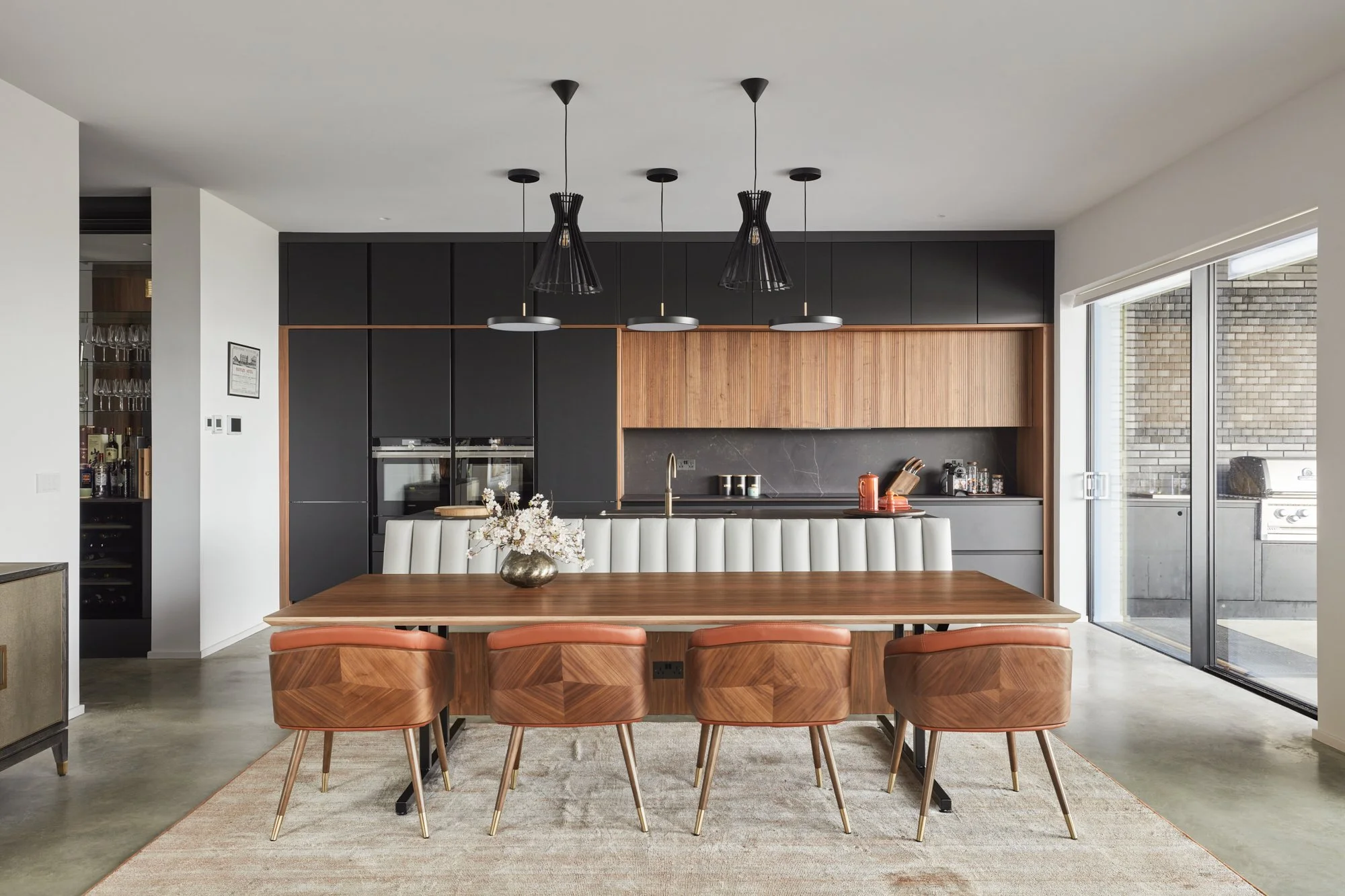 Modern kitchen with black and wood cabinets, a wooden dining table with tan chairs, pendant lighting, and sliding glass doors showing a brick wall outside.