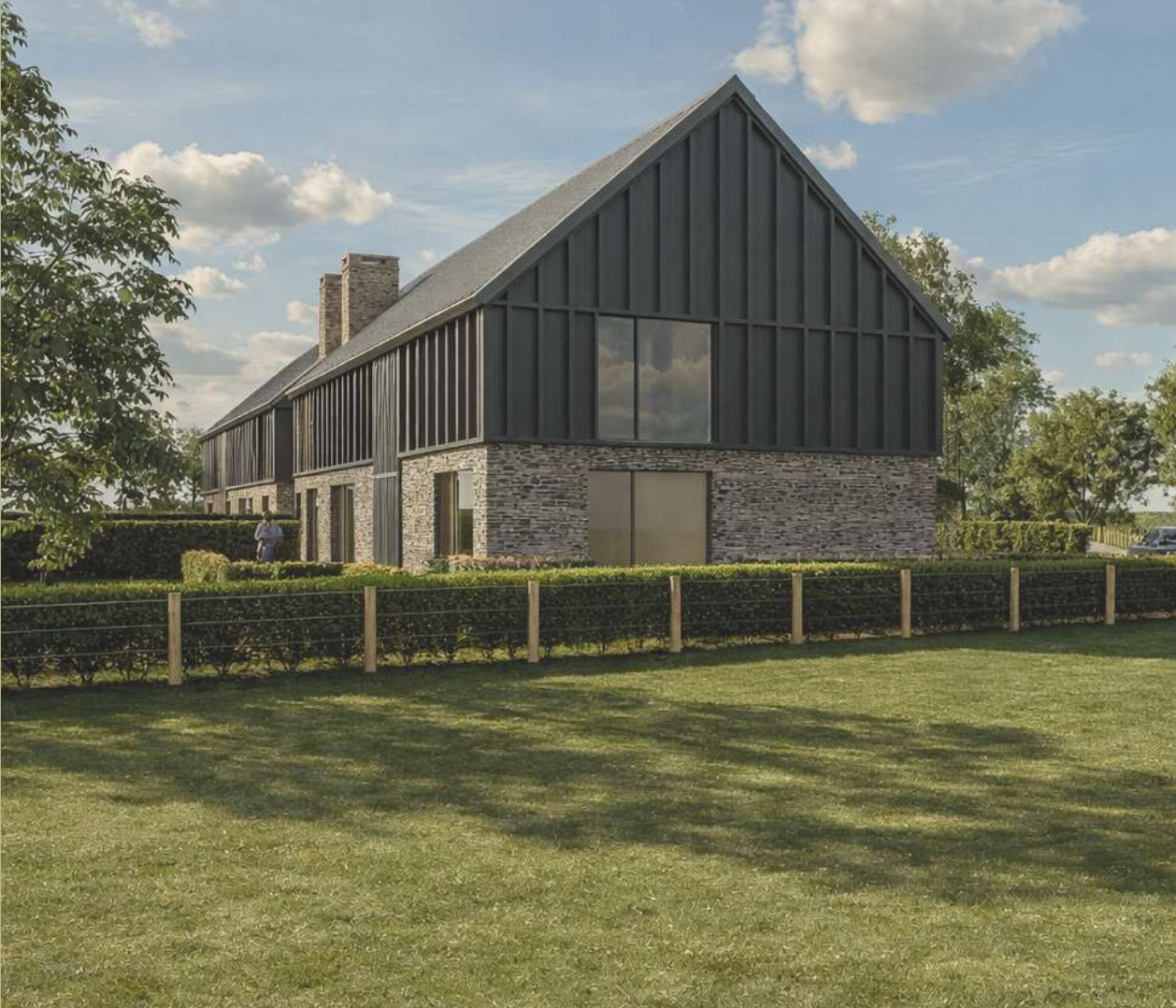Modern house with stone and black siding exterior, large sloped roof, and large surrounding lawn with a small wooden fence and trees in the background.