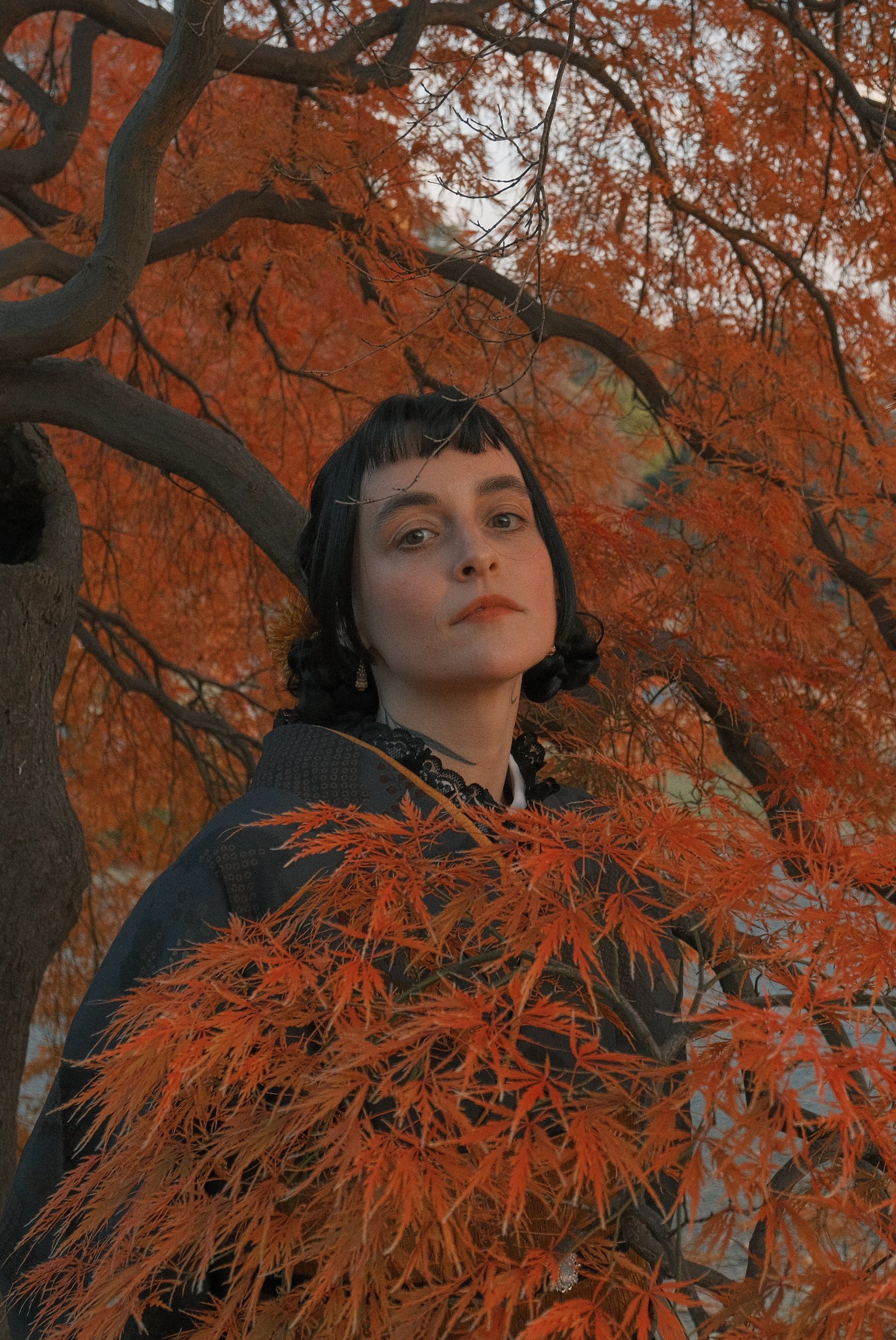 A woman with dark hair and pale skin stands behind orange autumn leaves, wearing a black garment with lace details, in front of a tree with orange foliage.