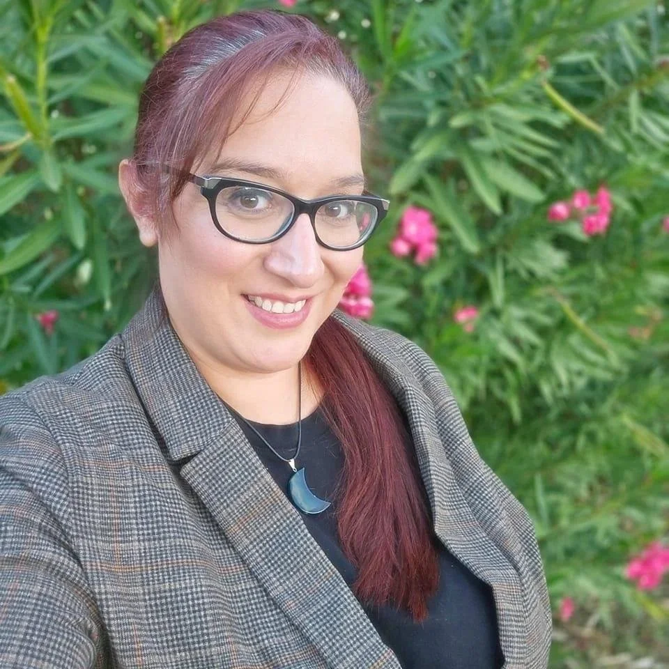 Kristen Gutierrez. A woman with glasses and reddish-brown hair taking a selfie outdoors, dressed in a plaid blazer and wearing a moon-shaped necklace, with pink flowers and green foliage in the background.