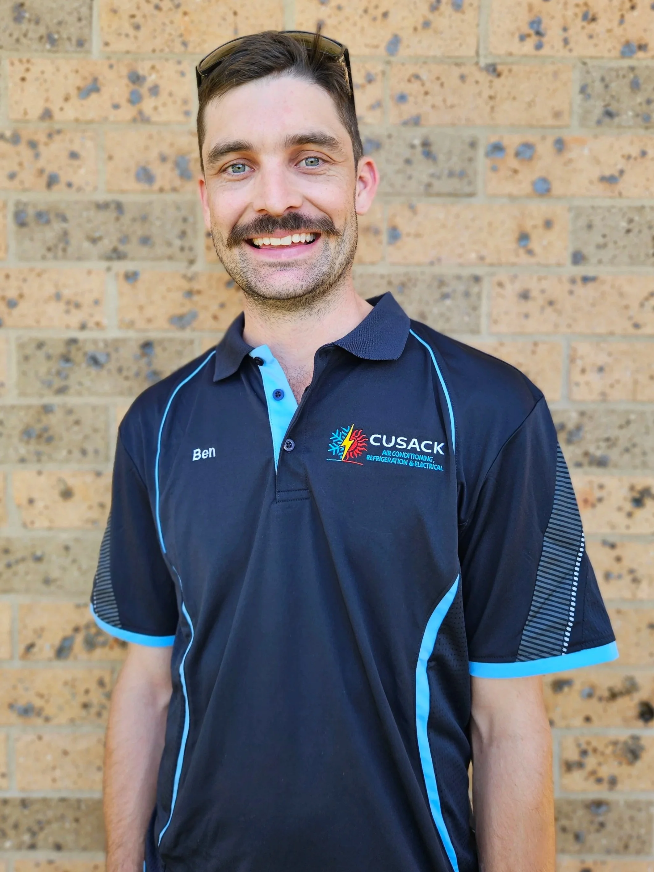 A man with short dark hair, blue eyes, and a beard, smiling and standing against a brick wall. He is wearing sunglasses on his head and a navy blue polo shirt with light blue accents and the logo of CUSACK Air Conditioning, Refrigeration & Electrical. The shirt has his name, Ben, embroidered on it.