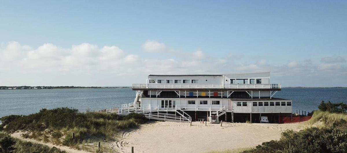 Beachside building with multiple levels, white exterior, surrounded by sand and dunes, with water and sky in the background.