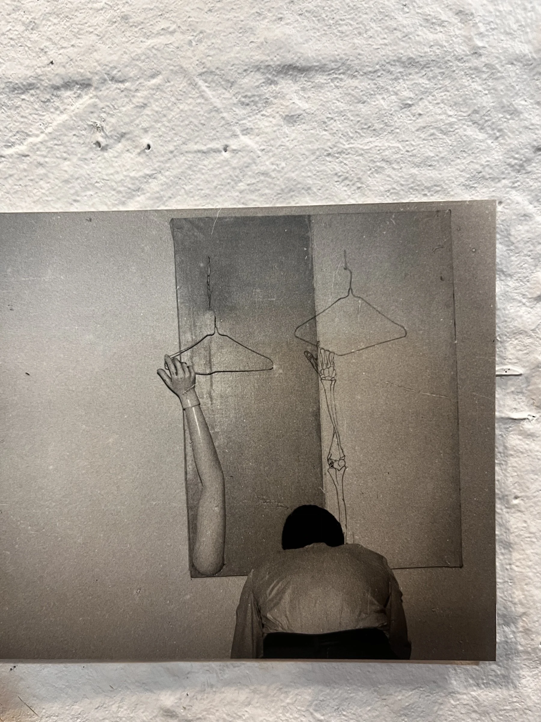 A black and white drawing of a human skeleton, with a highlighted arm and hand reaching to grab a coat hanger, on a wall with a small painted figure hanging from a string.