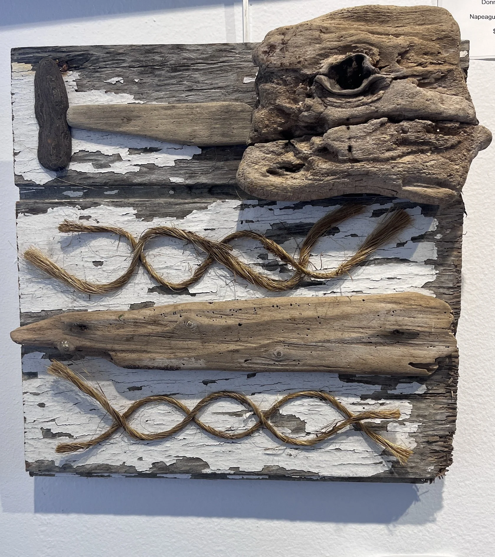 A display of three pieces of driftwood arranged on a distressed white wooden board. The top piece is weathered and rough, the middle piece is smoother, and the bottom piece is elongated with small holes. A twisted piece of rope is laid out in front of the driftwood.
